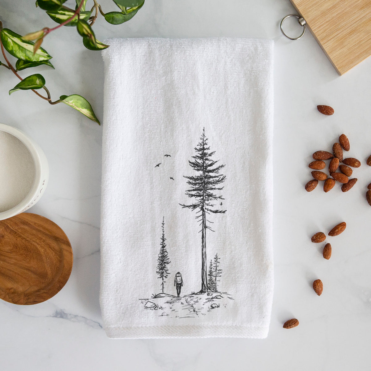 White hand towel printed with a black ink illustration of a lone hiker walking between tall pine trees with birds overhead.