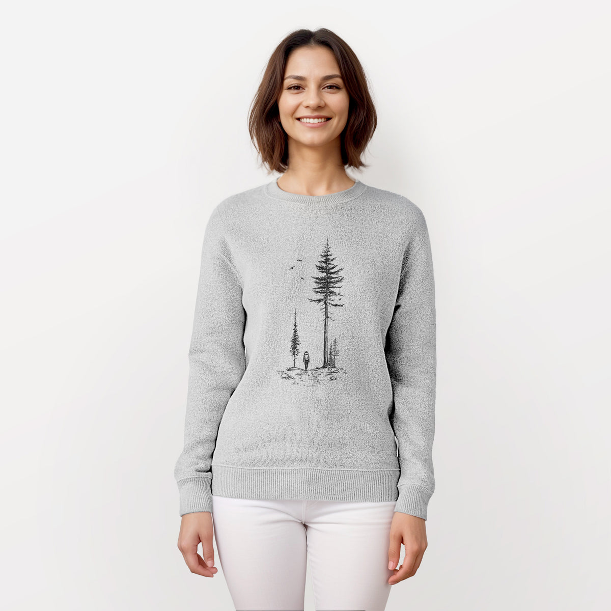 Woman wearing a knit sweatshirt printed with a black ink illustration of a lone hiker walking between tall pine trees with birds overhead.