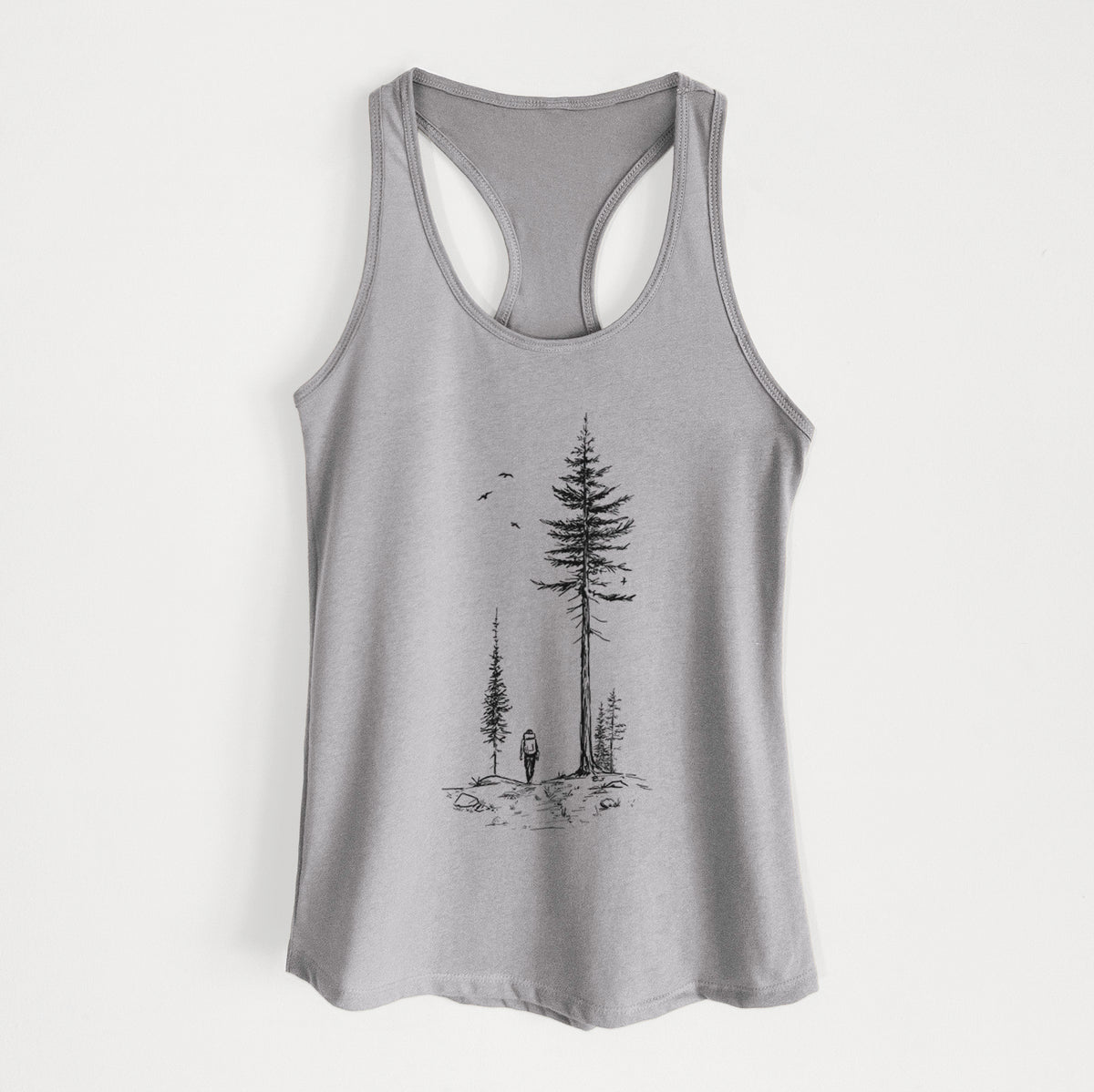 Heather grey women's racerback tank top laid flat, printed with a black ink illustration of a lone hiker walking between tall pine trees with birds overhead.