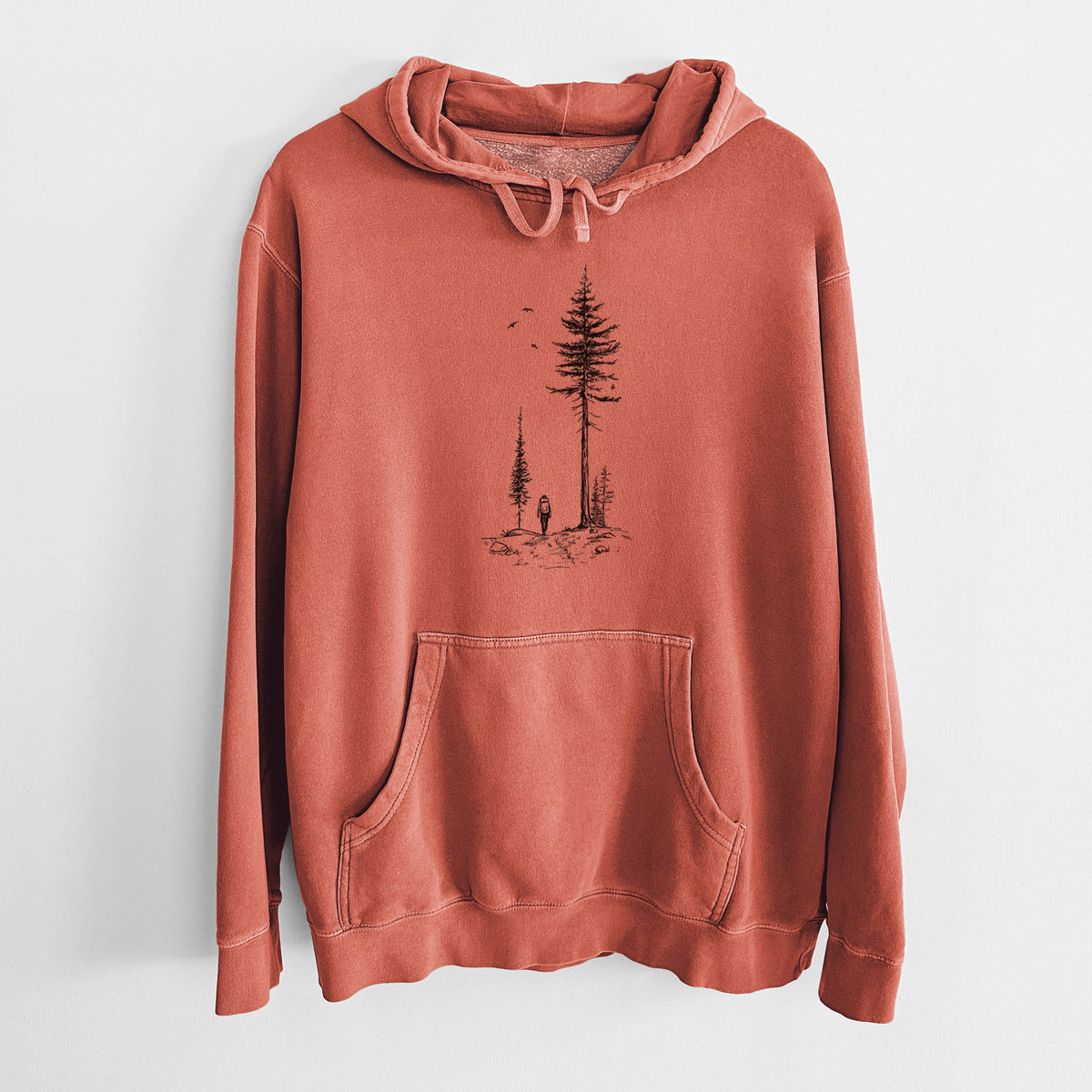 Amber orange pigment dyed hoodie laid flat, printed with a black ink illustration of a lone hiker walking between tall pine trees with birds overhead.