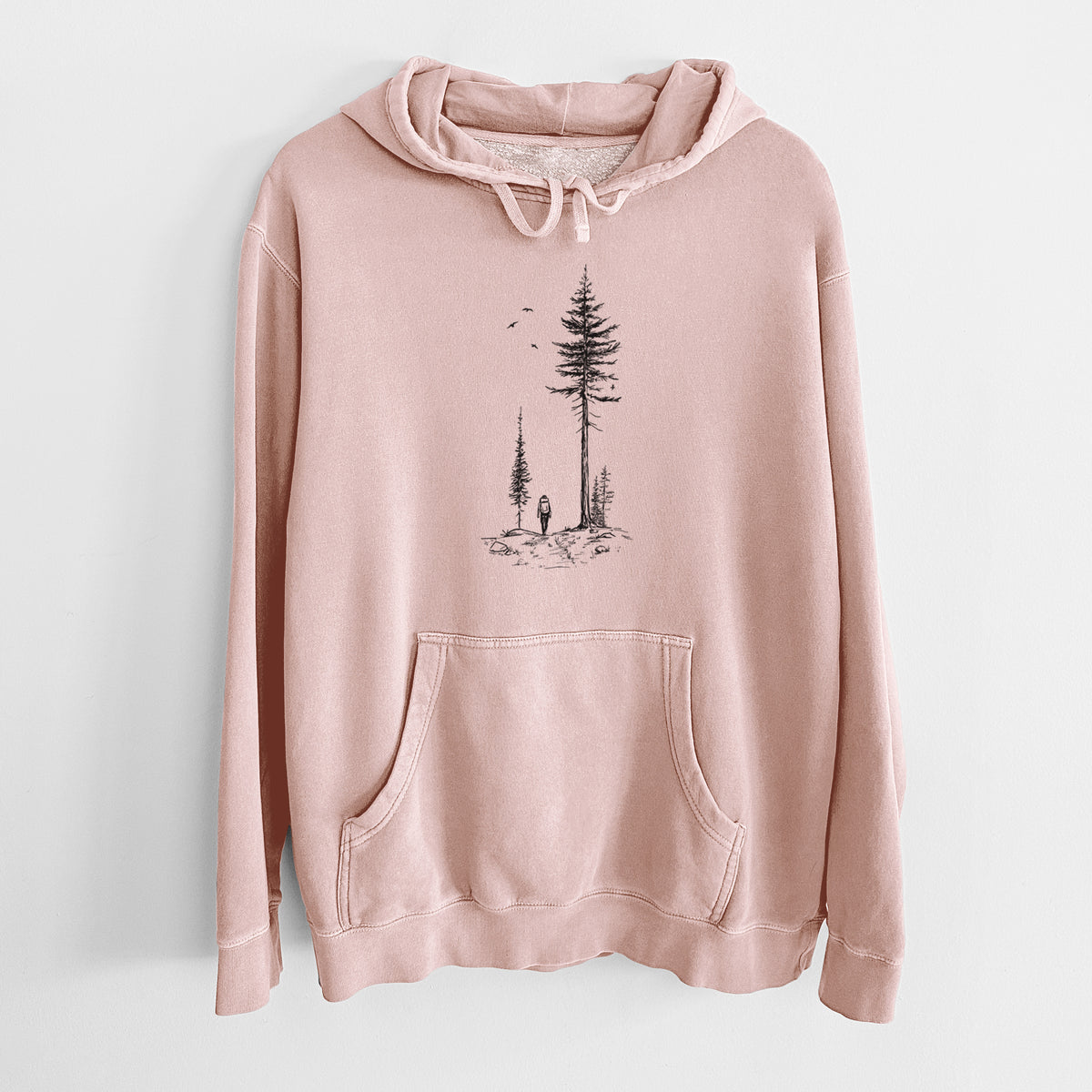 Dusty pink pigment dyed hoodie laid flat, printed with a black ink illustration of a lone hiker walking between tall pine trees with birds overhead.