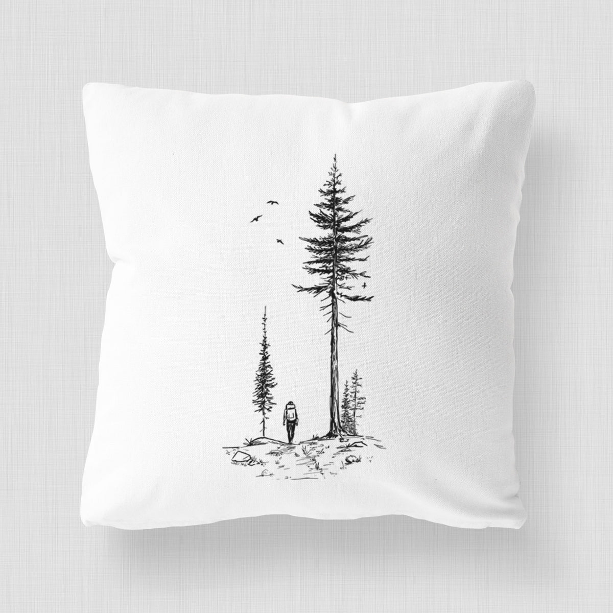 White pillow cover printed with a black ink illustration of a lone hiker walking between tall pine trees with birds overhead.