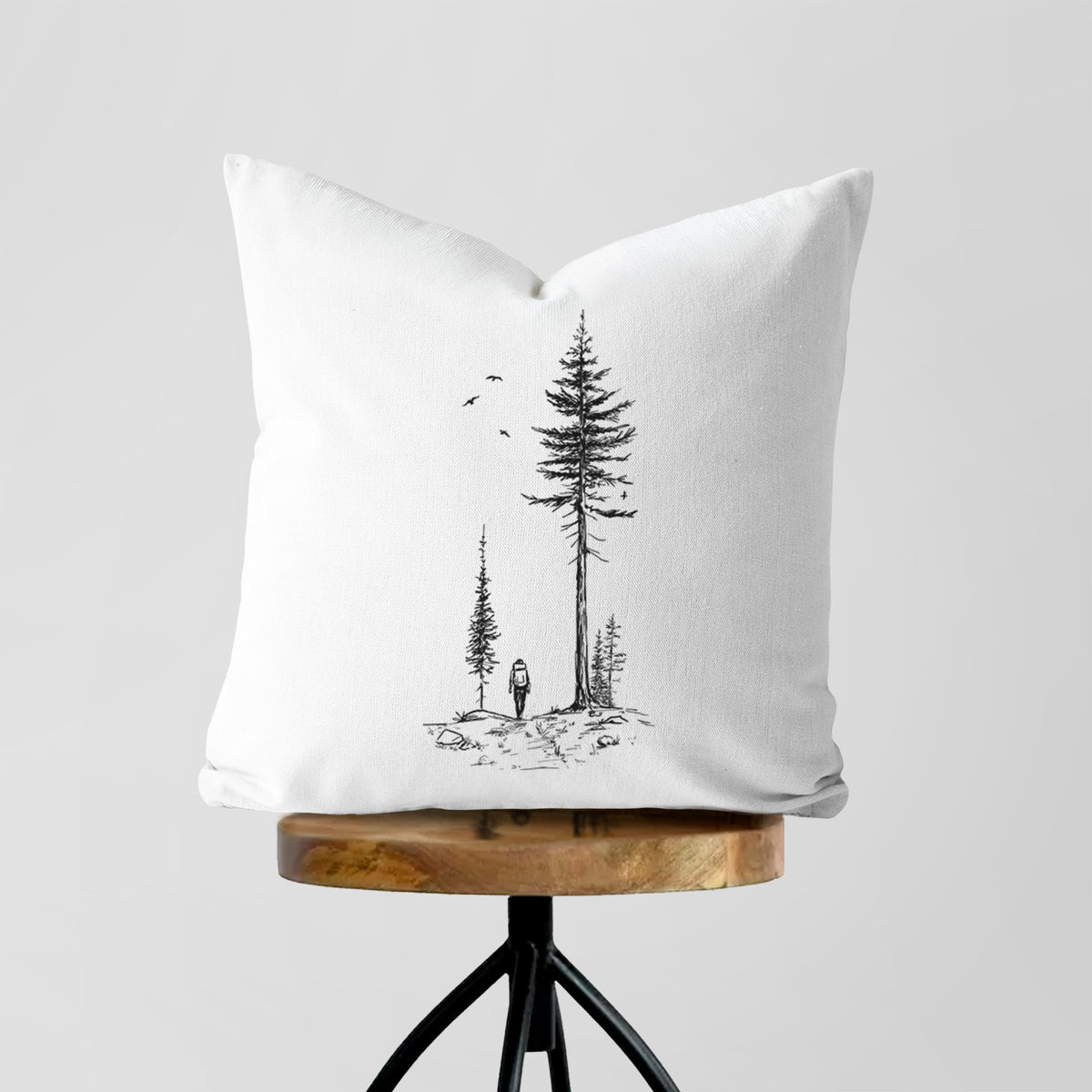 White pillow cover printed with a black ink illustration of a lone hiker walking between tall pine trees with birds overhead, on a wooden stool.