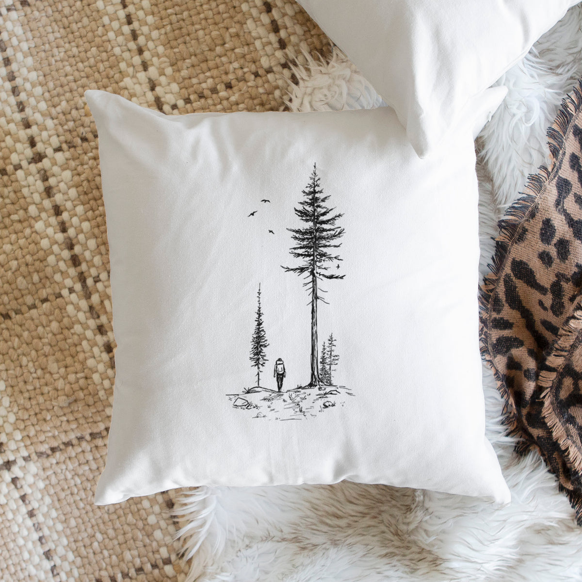 White pillow cover printed with a black ink illustration of a lone hiker walking between tall pine trees with birds overhead, on a woven chair.