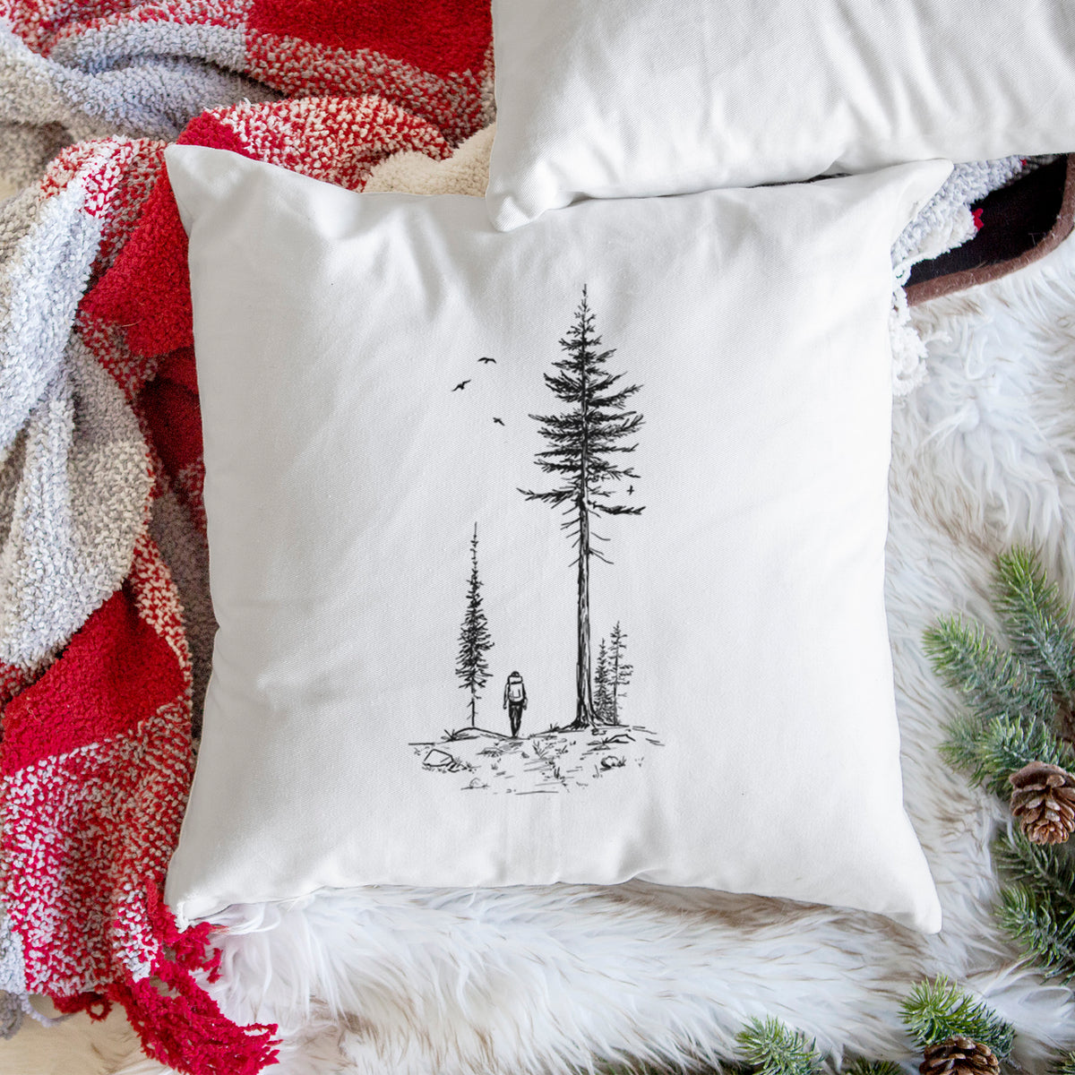 White pillow cover printed with a black ink illustration of a lone hiker walking between tall pine trees with birds overhead, on a woven chair.