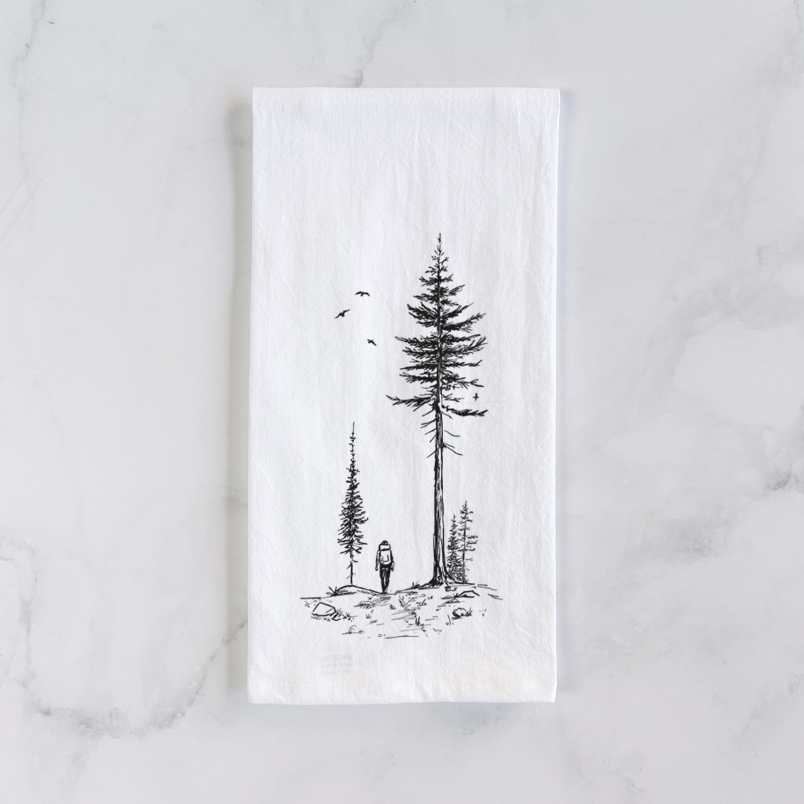 White cotton tea towel printed with a black ink illustration of a lone hiker walking between tall pine trees with birds overhead, on a marble counter beside a round wooden cutting board and greenery.