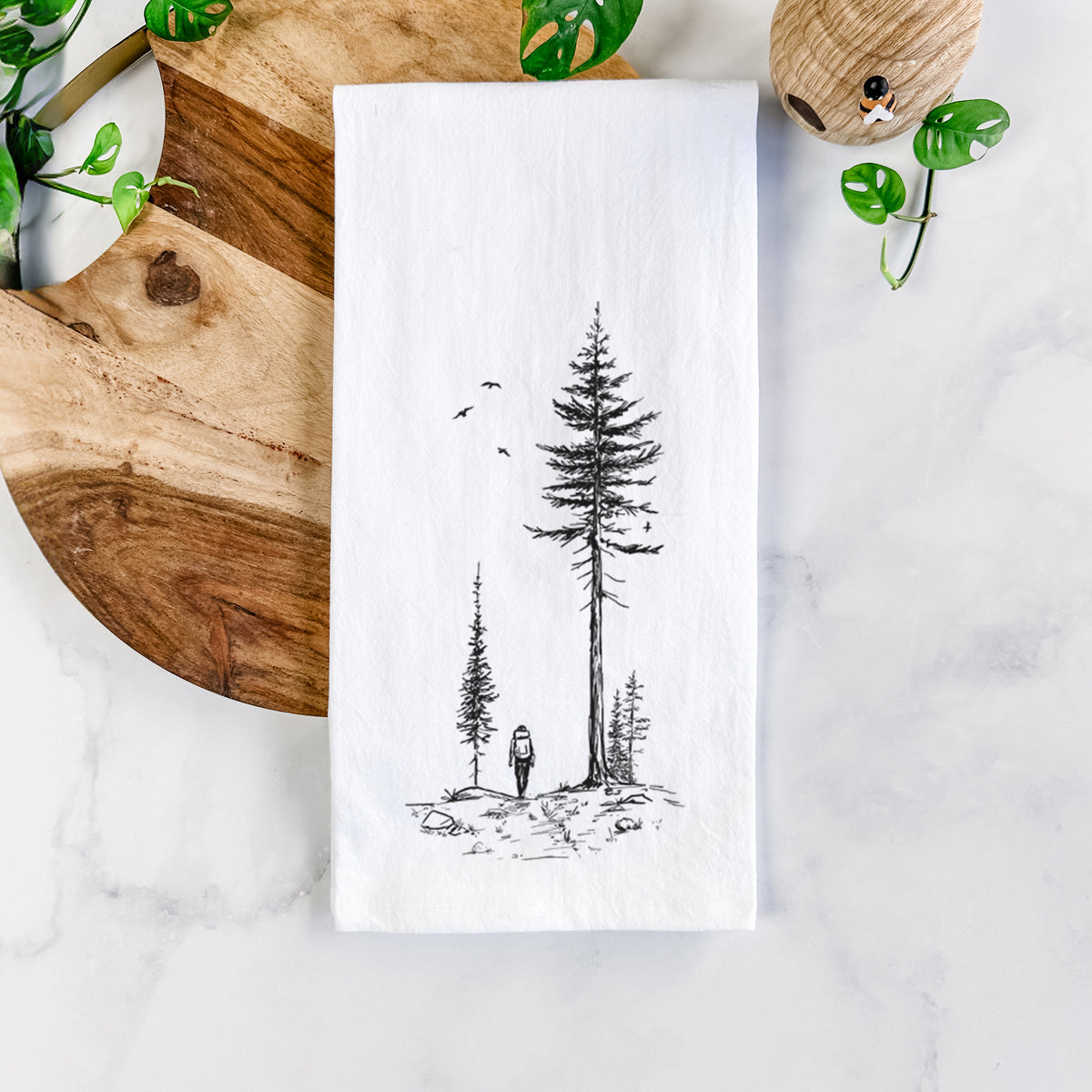 White cotton tea towel printed with a black ink illustration of a lone hiker walking between tall pine trees with birds overhead, on a marble counter beside a round wooden cutting board and greenery.
