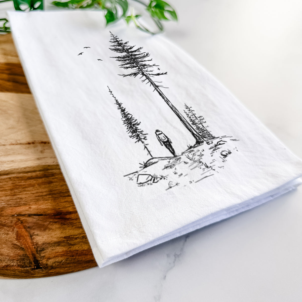 Close-up of a folded white tea towel on a wooden board, showing a black ink illustration of a lone hiker walking between tall pine trees with birds overhead.