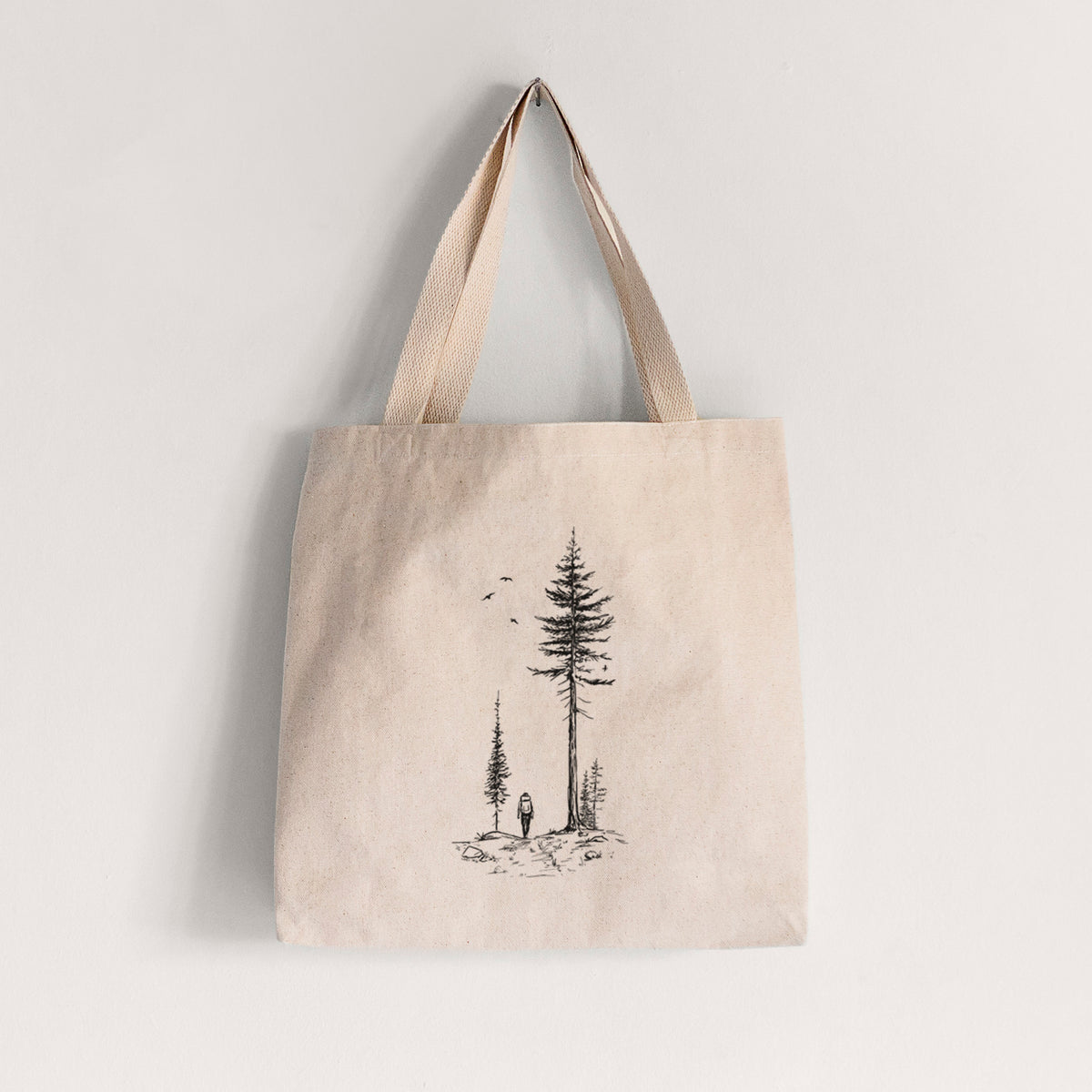 Natural canvas tote bag printed with a black ink illustration of a lone hiker walking between tall pine trees with birds overhead.