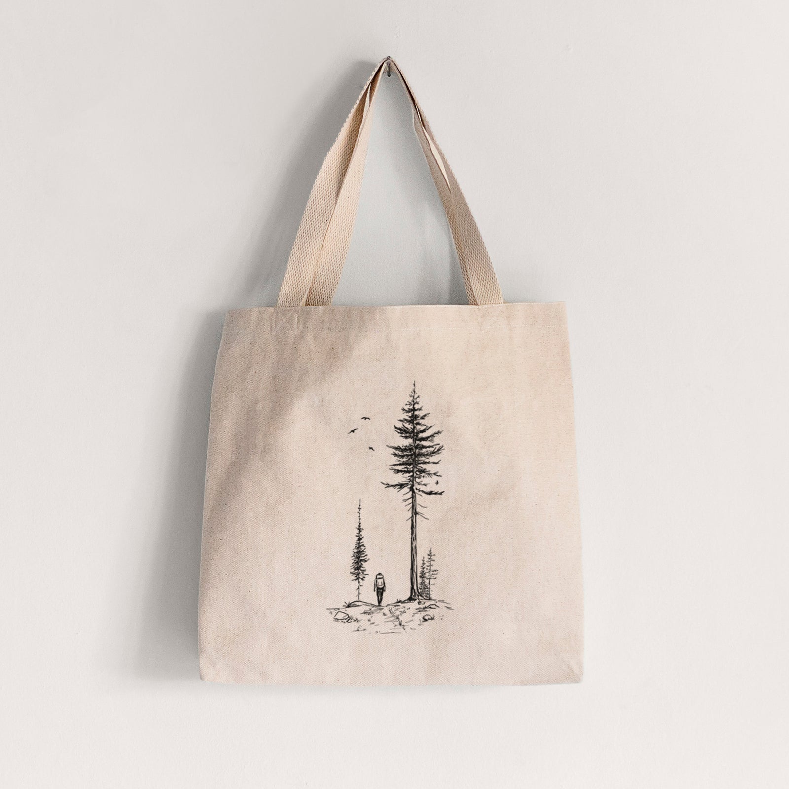Natural canvas tote bag printed with a black ink illustration of a lone hiker walking between tall pine trees with birds overhead.