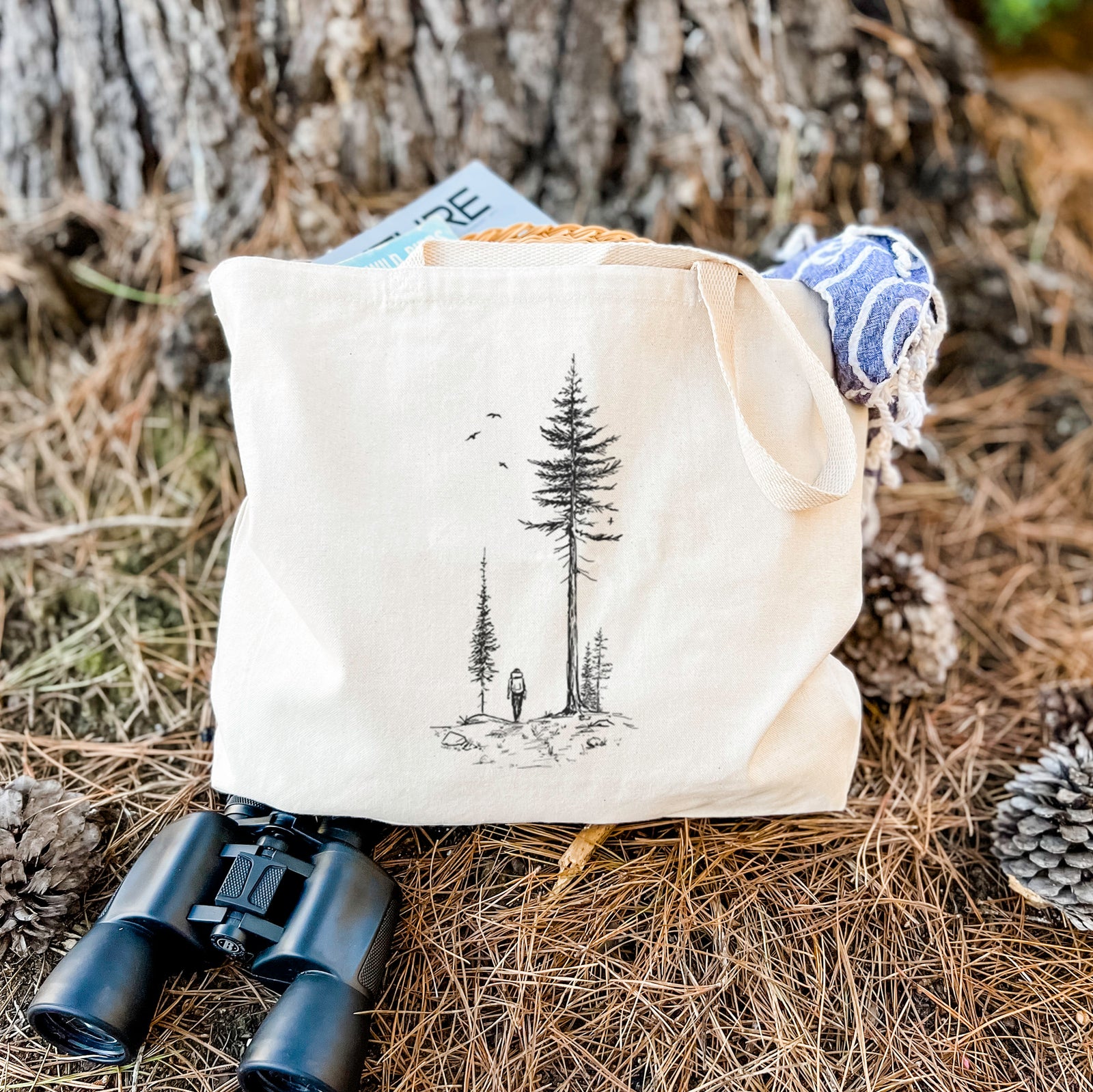 Natural canvas tote bag printed with a black ink illustration of a lone hiker walking between tall pine trees with birds overhead.