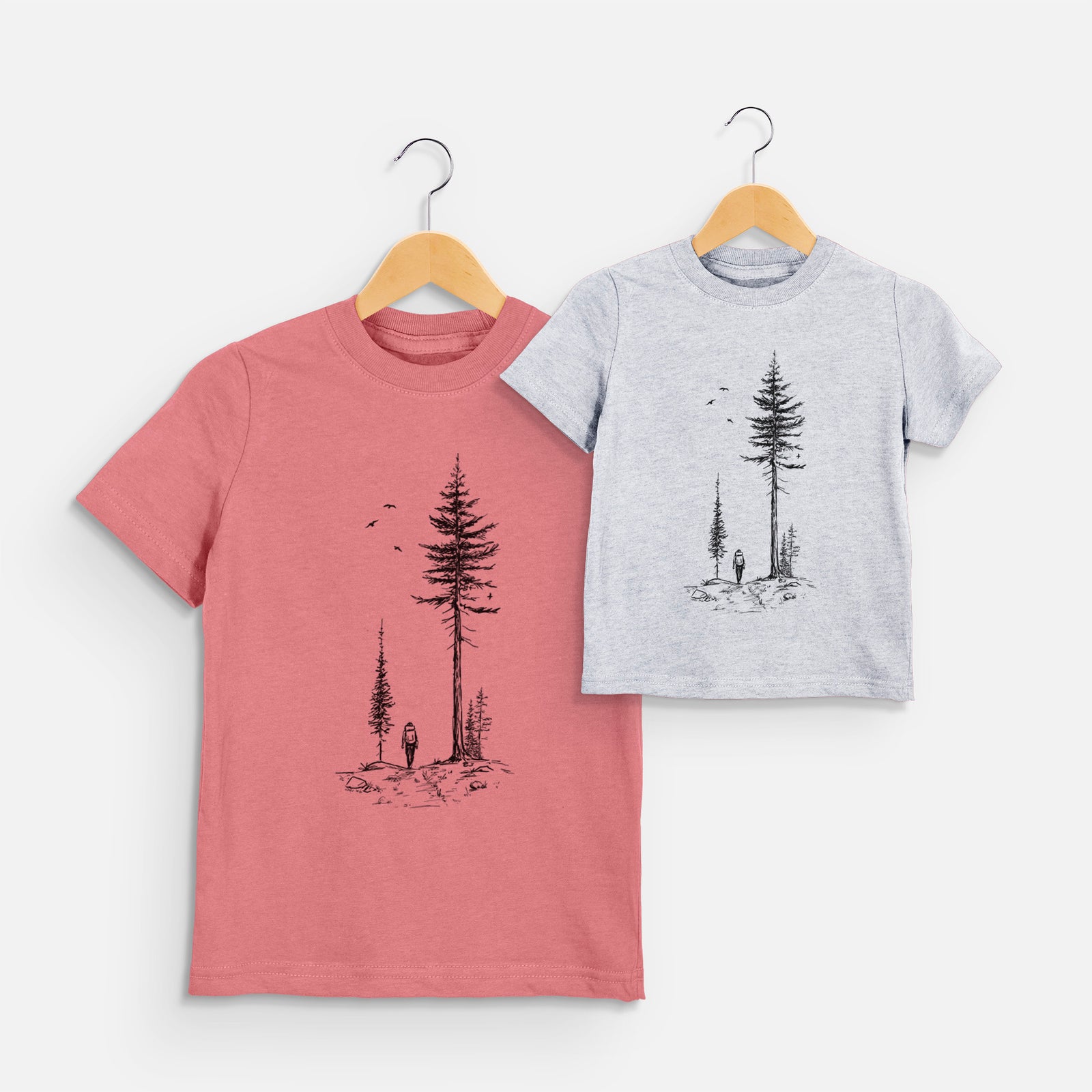 Two youth t-shirts in different colors on hangers, printed with a black ink illustration of a lone hiker walking between tall pine trees with birds overhead.