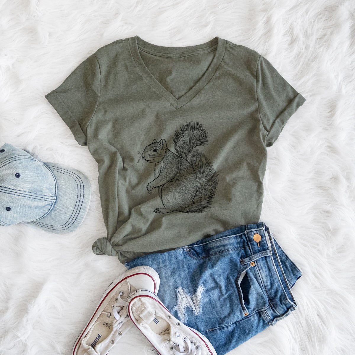 Military green women's relaxed fit cotton V-neck t-shirt printed with a detailed black ink illustration of a fox squirrel sitting with a bushy tail, styled flat on a white faux fur rug with a cap, jeans and sneakers.