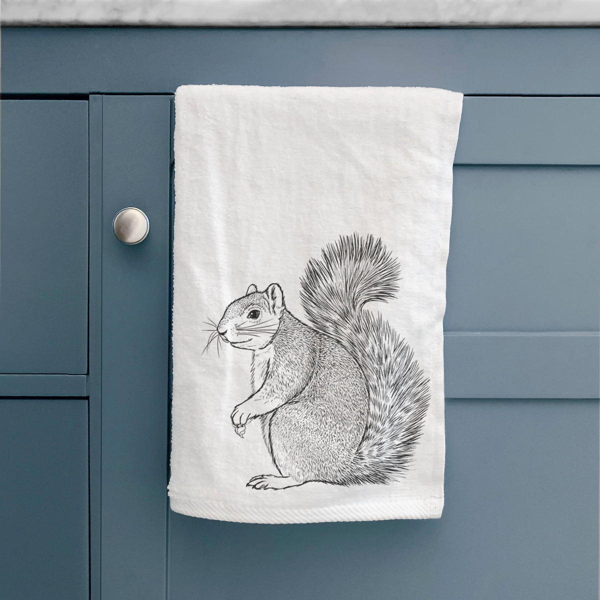 White hand towel printed with a detailed black ink illustration of a fox squirrel sitting with a bushy tail, hanging on a cabinet door.