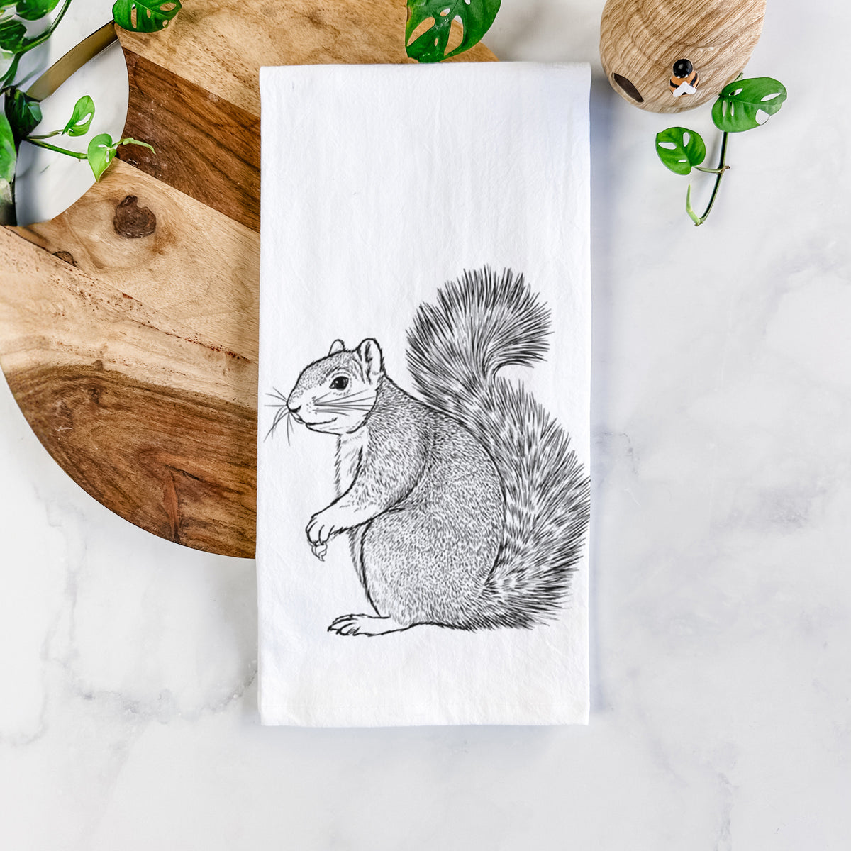 White cotton tea towel printed with a detailed black ink illustration of a fox squirrel sitting with a bushy tail, on a marble counter beside a round wooden cutting board and greenery.
