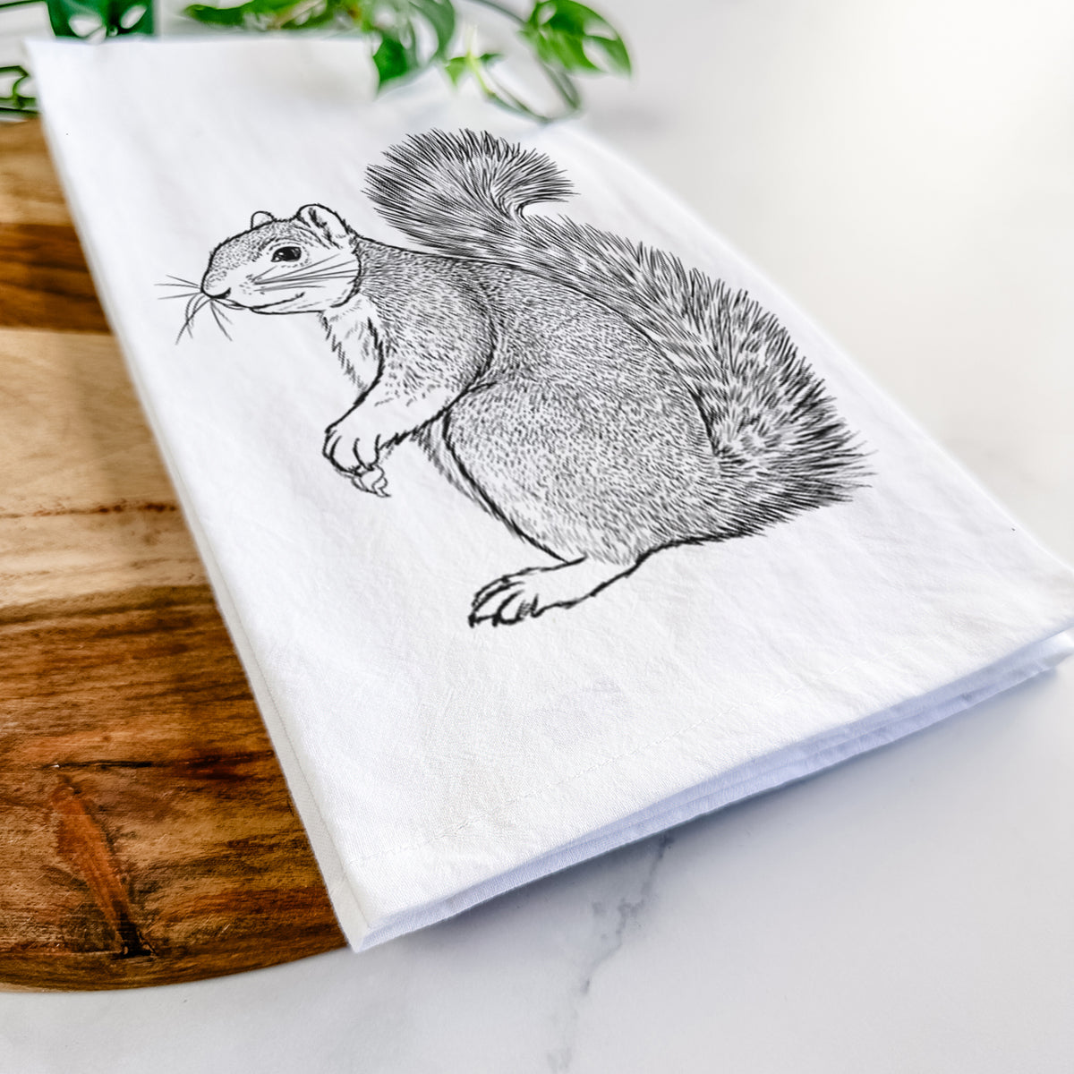 Close-up of a folded white tea towel on a wooden board, showing a detailed black ink illustration of a fox squirrel sitting with a bushy tail.