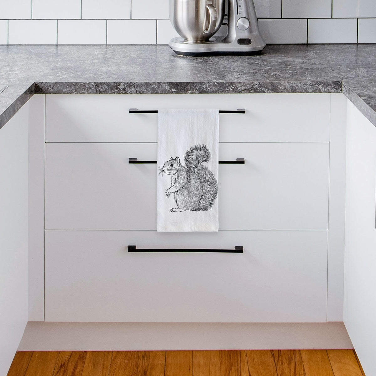 White tea towel printed with a detailed black ink illustration of a fox squirrel sitting with a bushy tail, hanging from a drawer handle in a bright white kitchen.