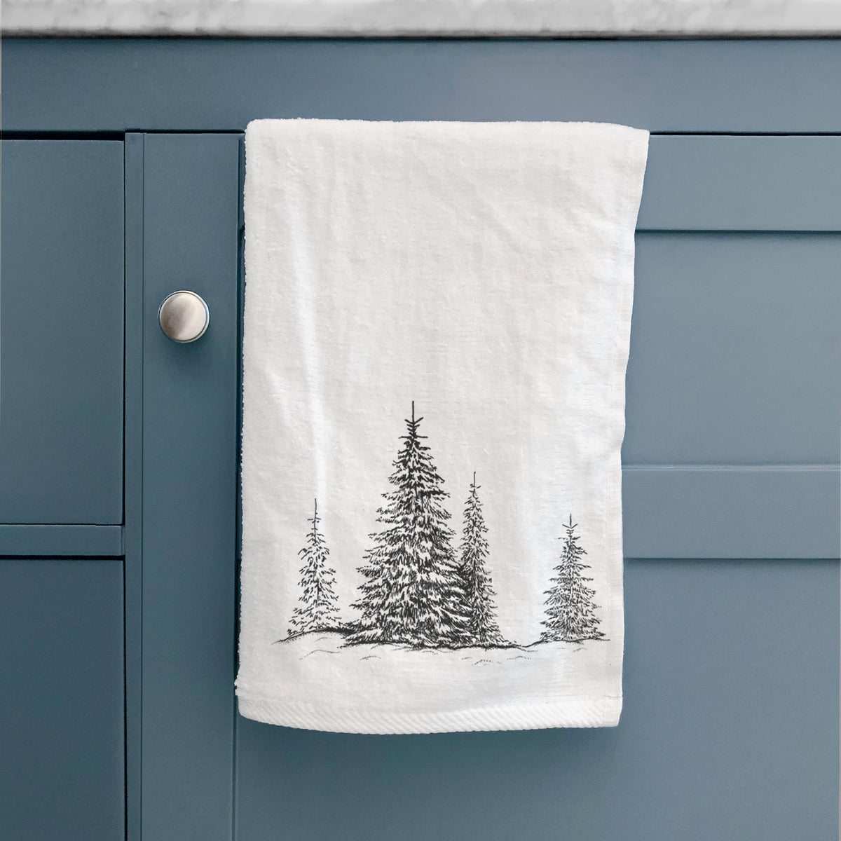 White hand towel printed with a black ink illustration of frost-covered evergreen trees, hanging on a cabinet door.