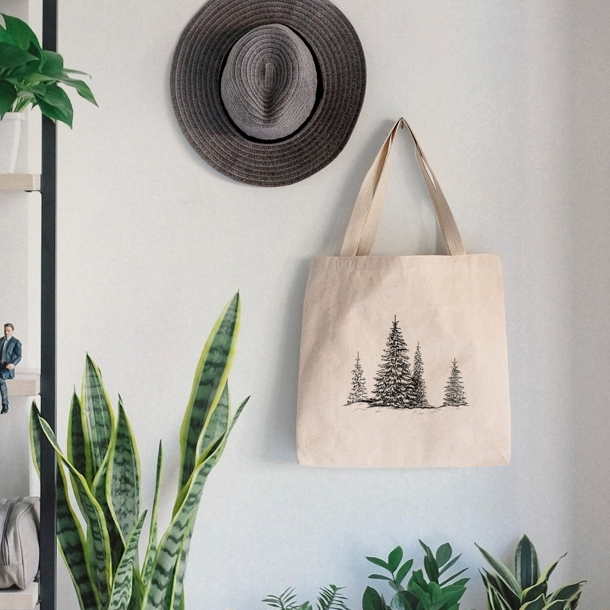 Canvas tote bag printed with a black ink illustration of frost-covered evergreen trees, hanging on a wall hook below a hat, next to potted plants.