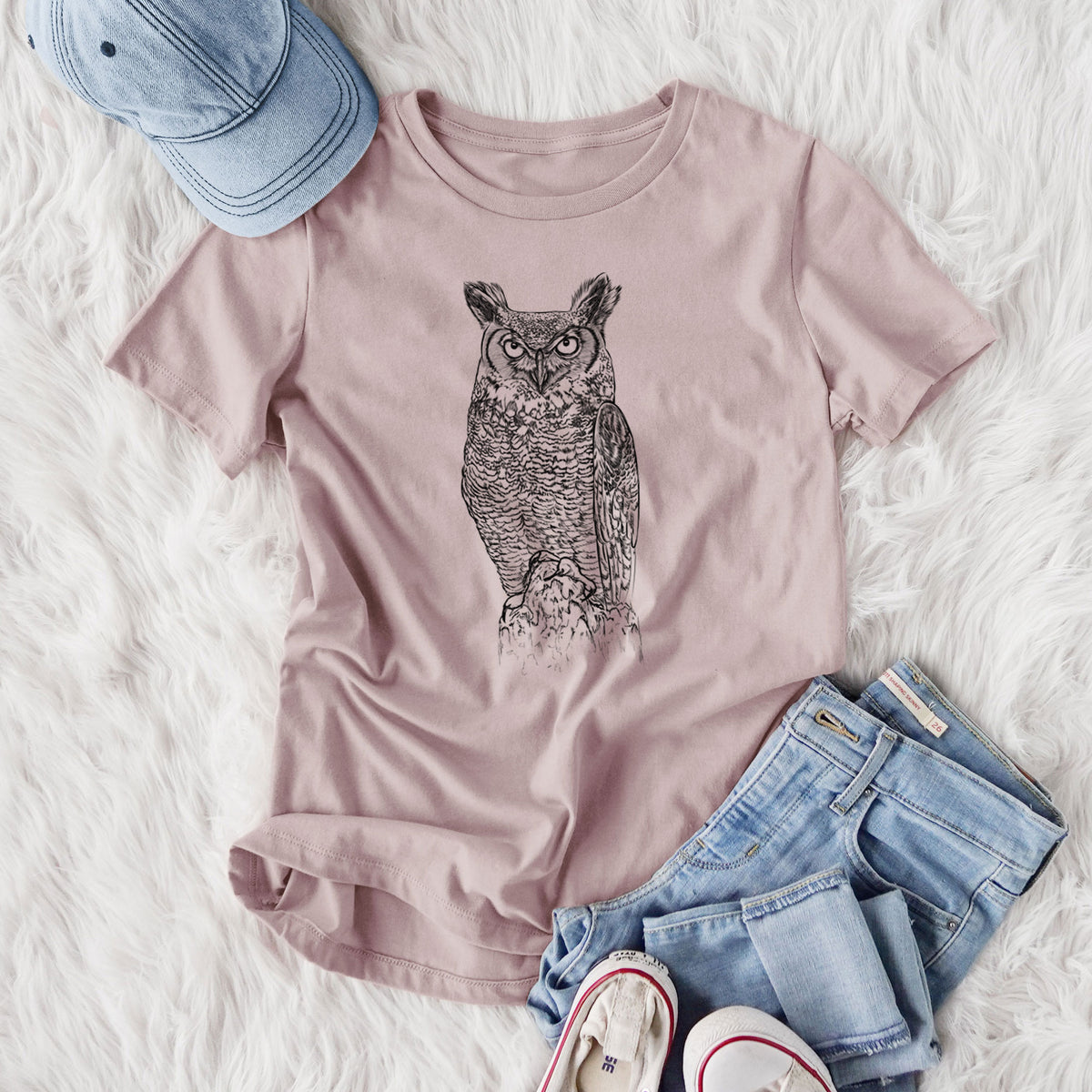 Women's relaxed fit cotton t-shirt printed with a detailed black ink illustration of a great horned owl standing upright with its ear tufts raised, styled flat on a white faux fur rug with jeans.