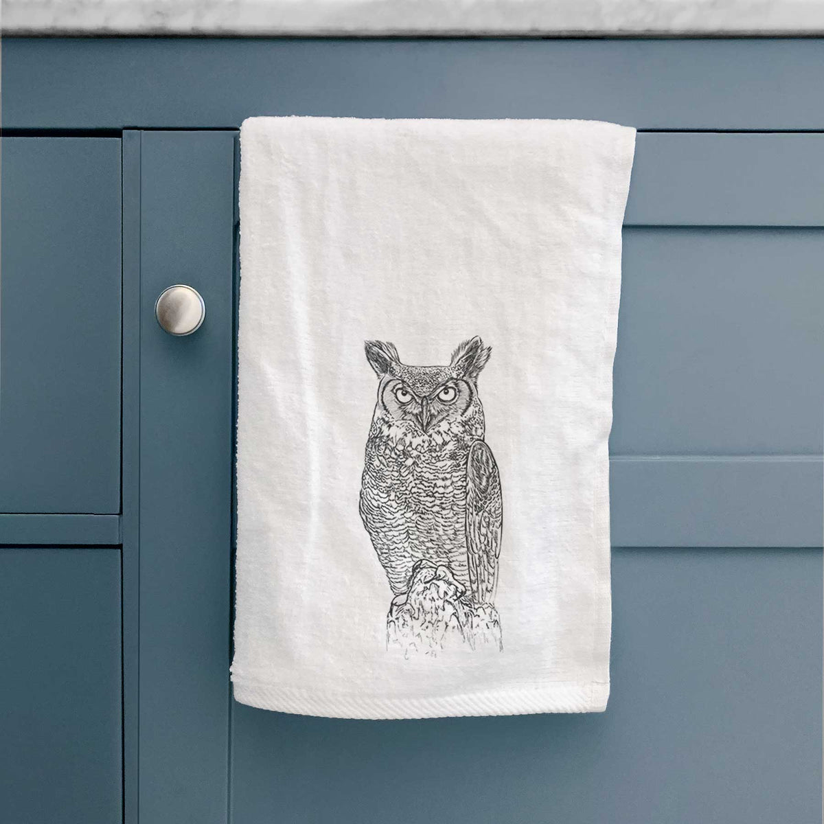 White hand towel printed with a detailed black ink illustration of a great horned owl standing upright with its ear tufts raised, hanging on a cabinet door.