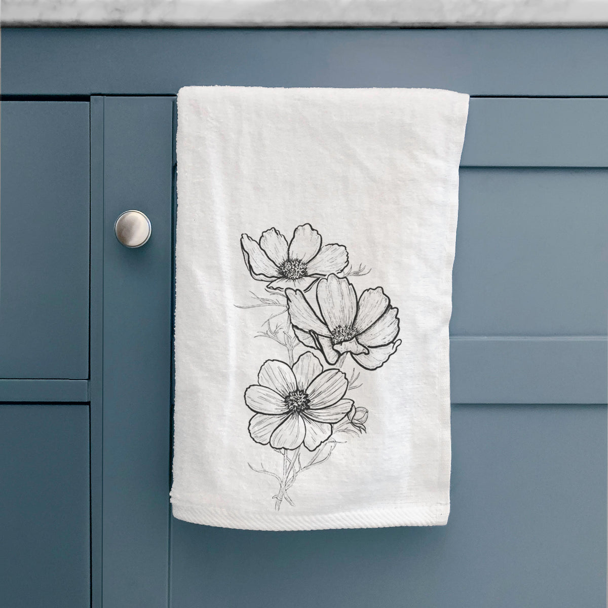 White hand towel printed with a black ink line drawing of garden cosmos flowers on stems, hanging on a cabinet door.