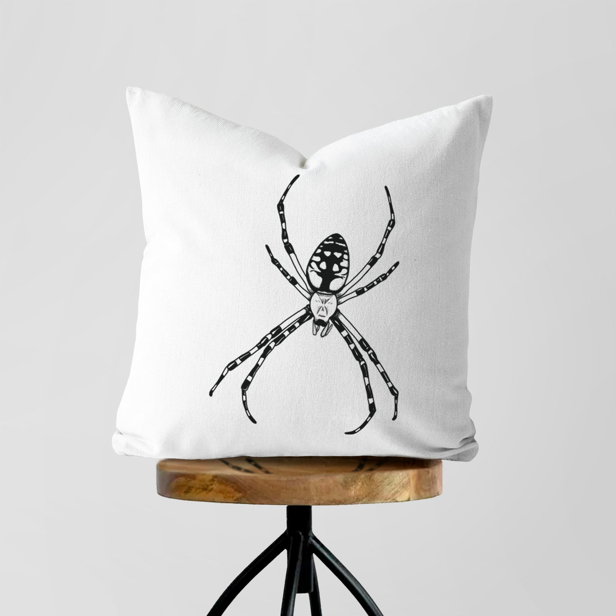 White pillow cover printed with a detailed black ink illustration of a garden spider with long striped legs, on a wooden stool.