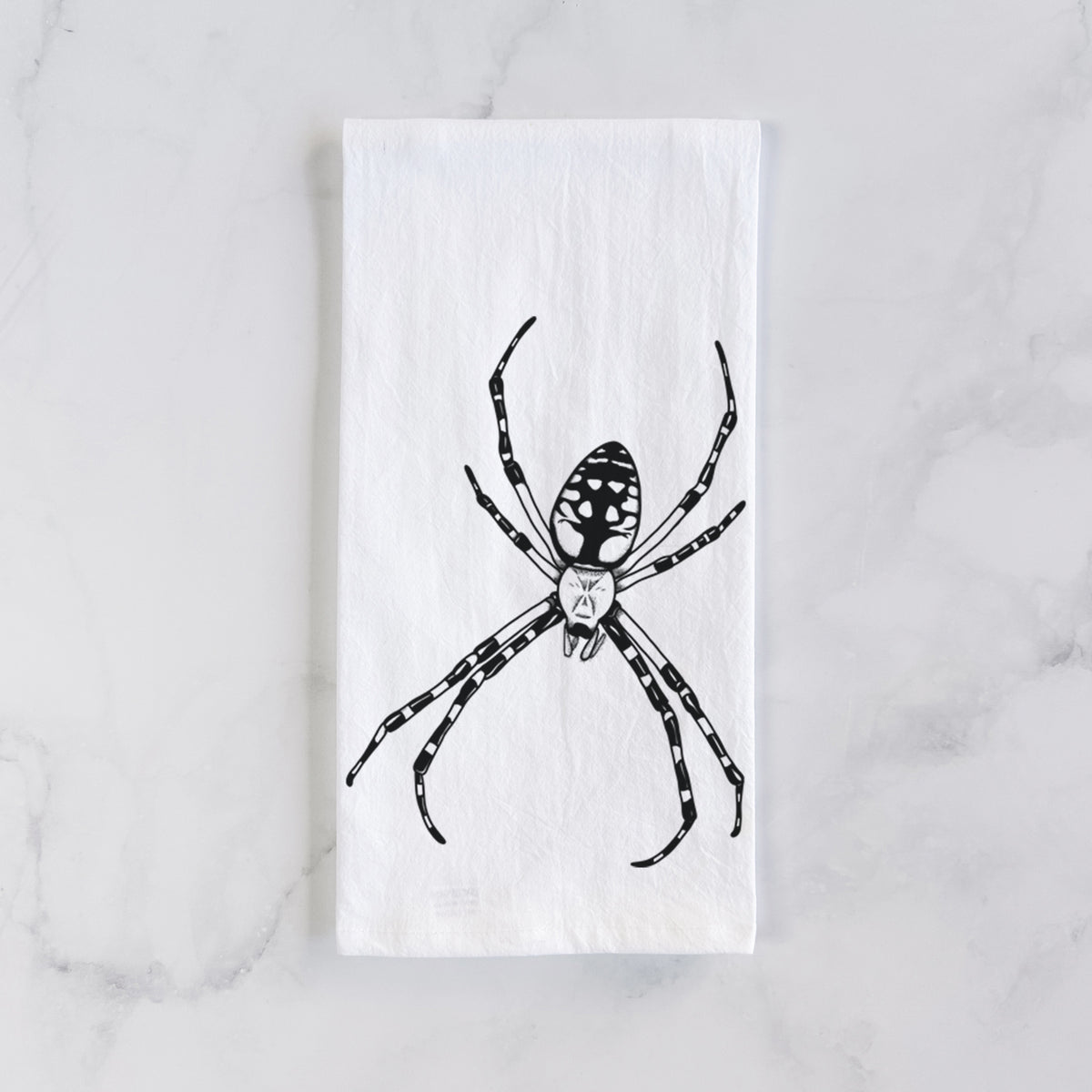 White cotton tea towel laid flat on marble, printed with a detailed black ink illustration of a garden spider with long striped legs.