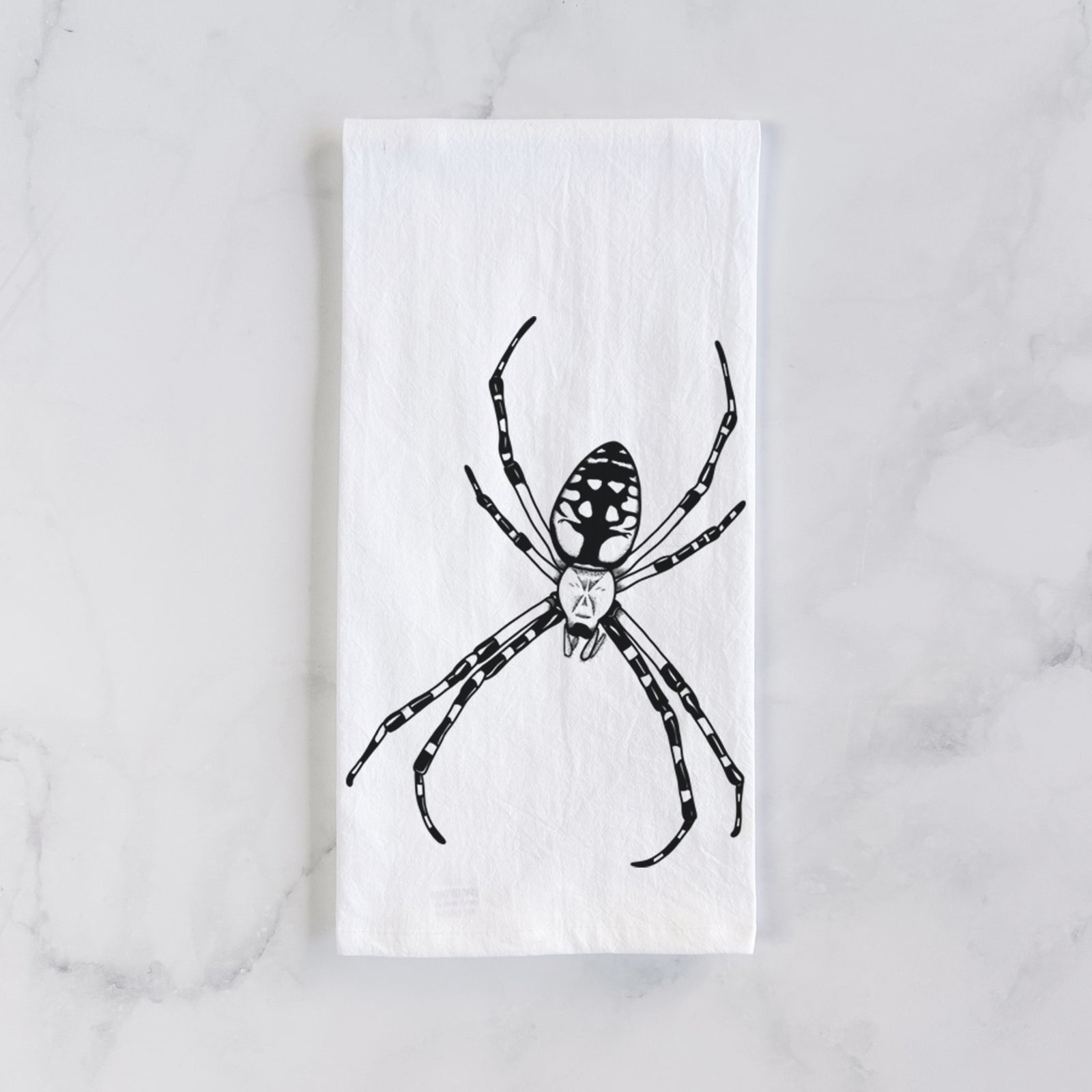 White cotton tea towel printed with a detailed black ink illustration of a garden spider with long striped legs, on a marble counter beside a round wooden cutting board and greenery.