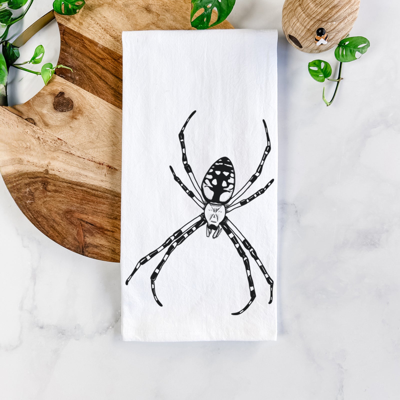White cotton tea towel printed with a detailed black ink illustration of a garden spider with long striped legs, on a marble counter beside a round wooden cutting board and greenery.