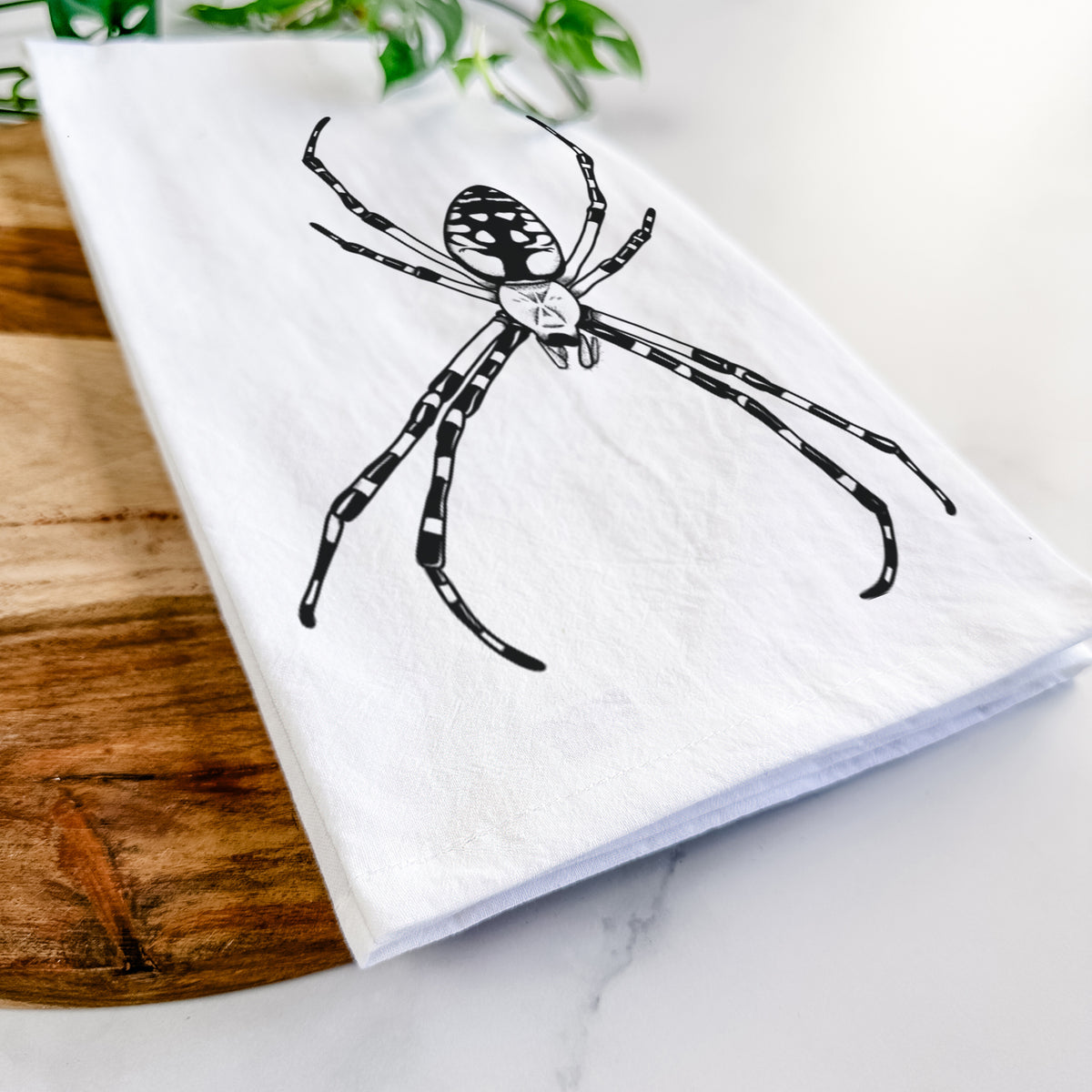 Close-up of a folded white tea towel on a wooden board, showing a detailed black ink illustration of a garden spider with long striped legs.