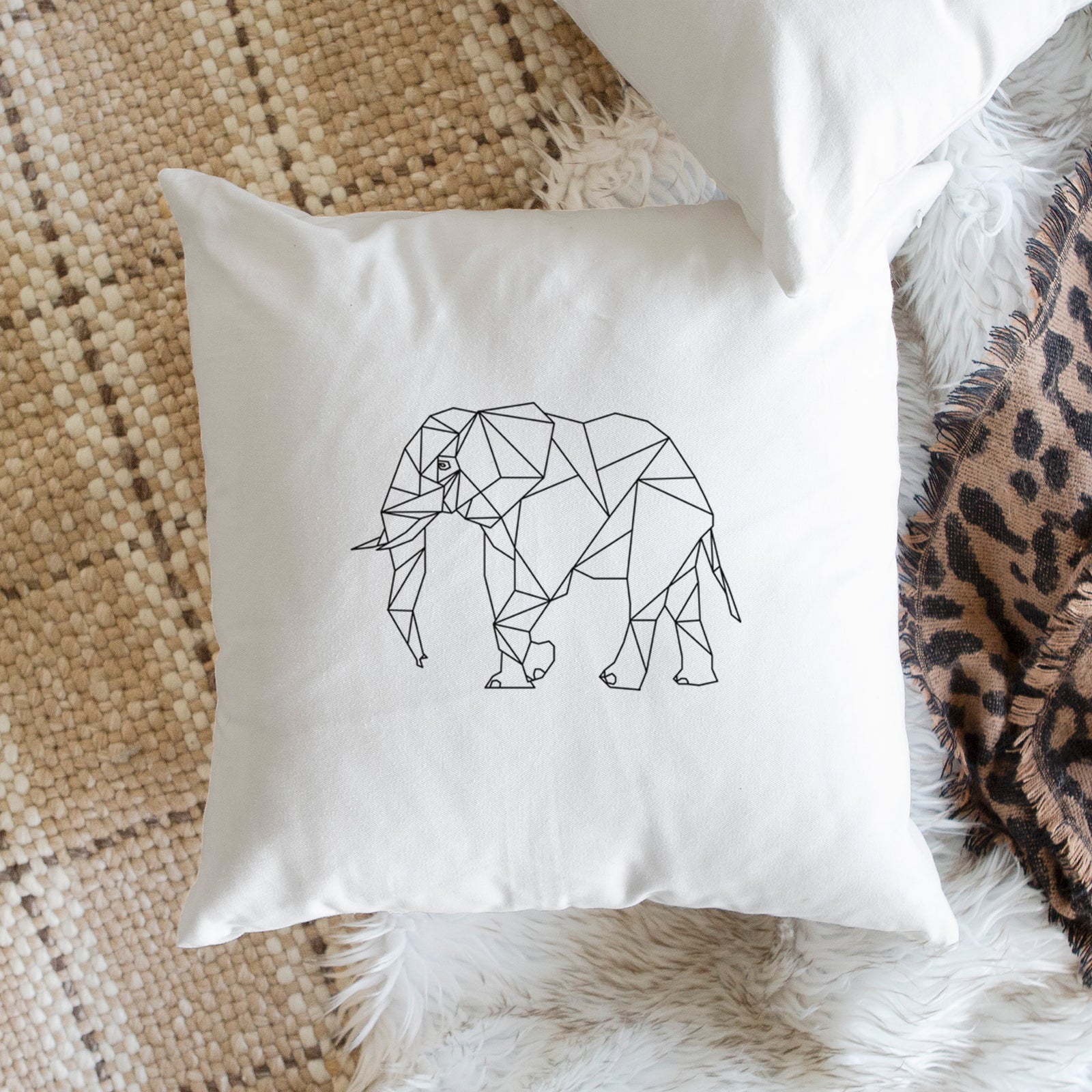 White pillow cover printed with a black geometric line art elephant, on a bench beside plants.