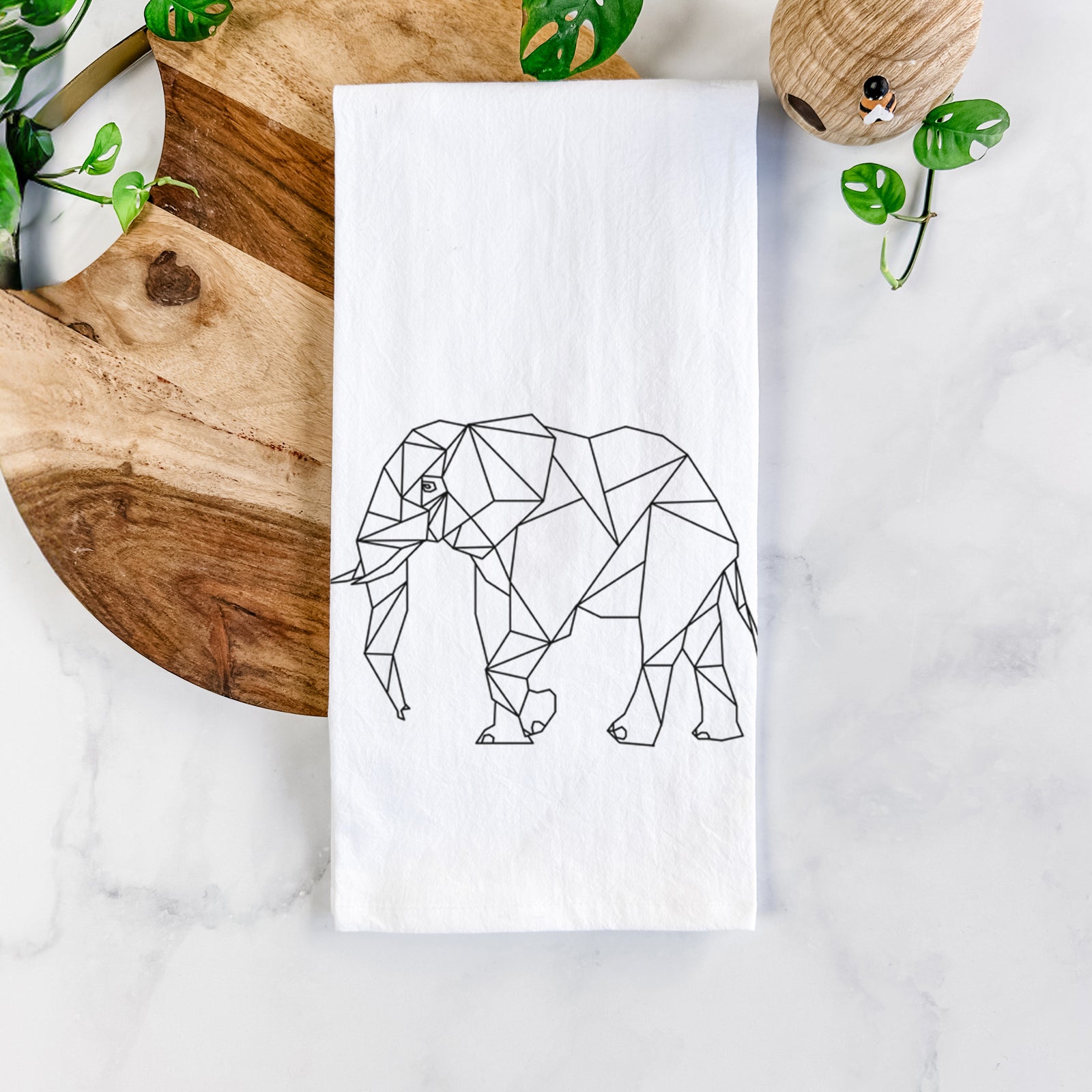White cotton tea towel printed with a black geometric line art elephant, on a marble counter beside a round wooden cutting board and greenery.