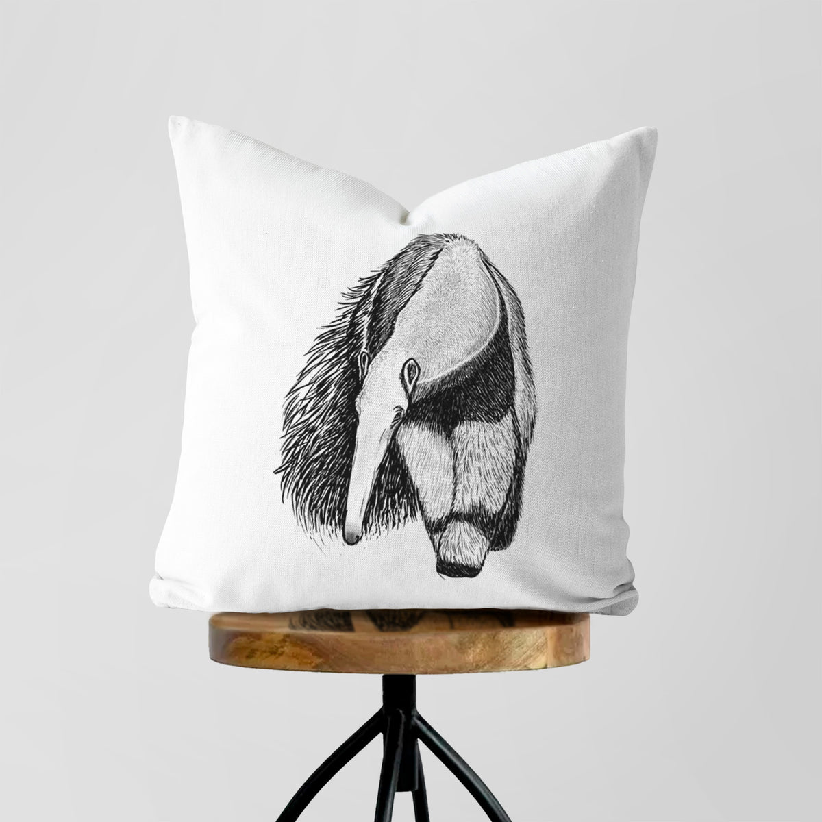 White pillow cover printed with a detailed black ink illustration of a giant anteater with its long snout and bushy tail, on a wooden stool.