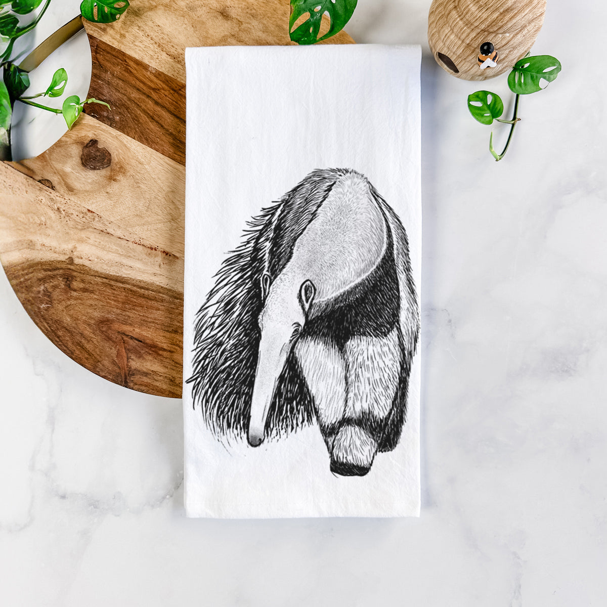 White cotton tea towel printed with a detailed black ink illustration of a giant anteater with its long snout and bushy tail, on a marble counter beside a round wooden cutting board and greenery.