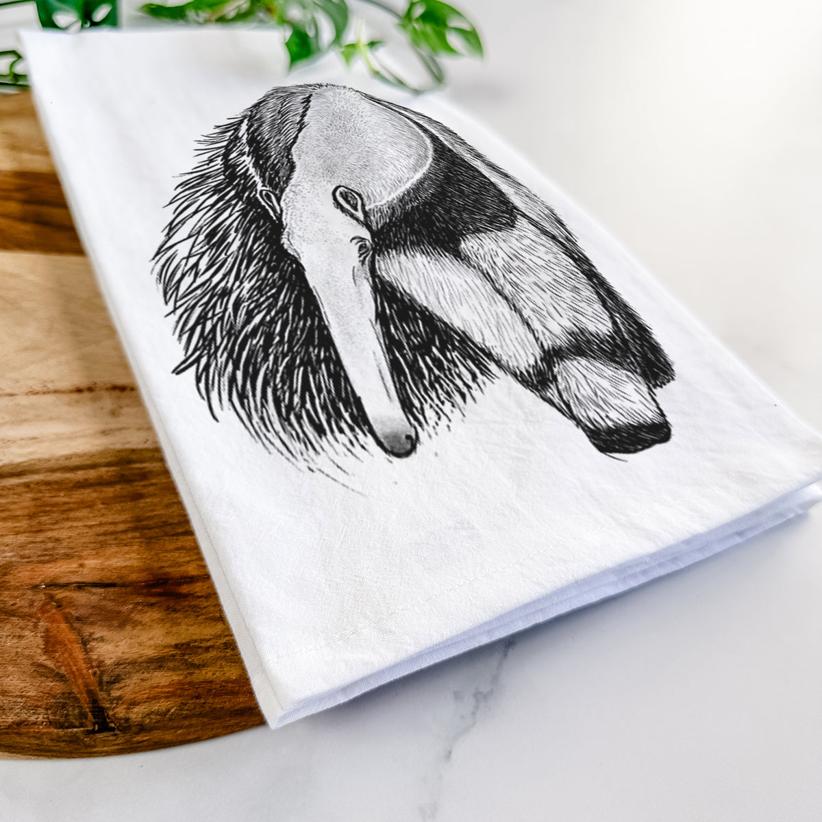 Close-up of a folded white tea towel on a wooden board, showing a detailed black ink illustration of a giant anteater with its long snout and bushy tail.