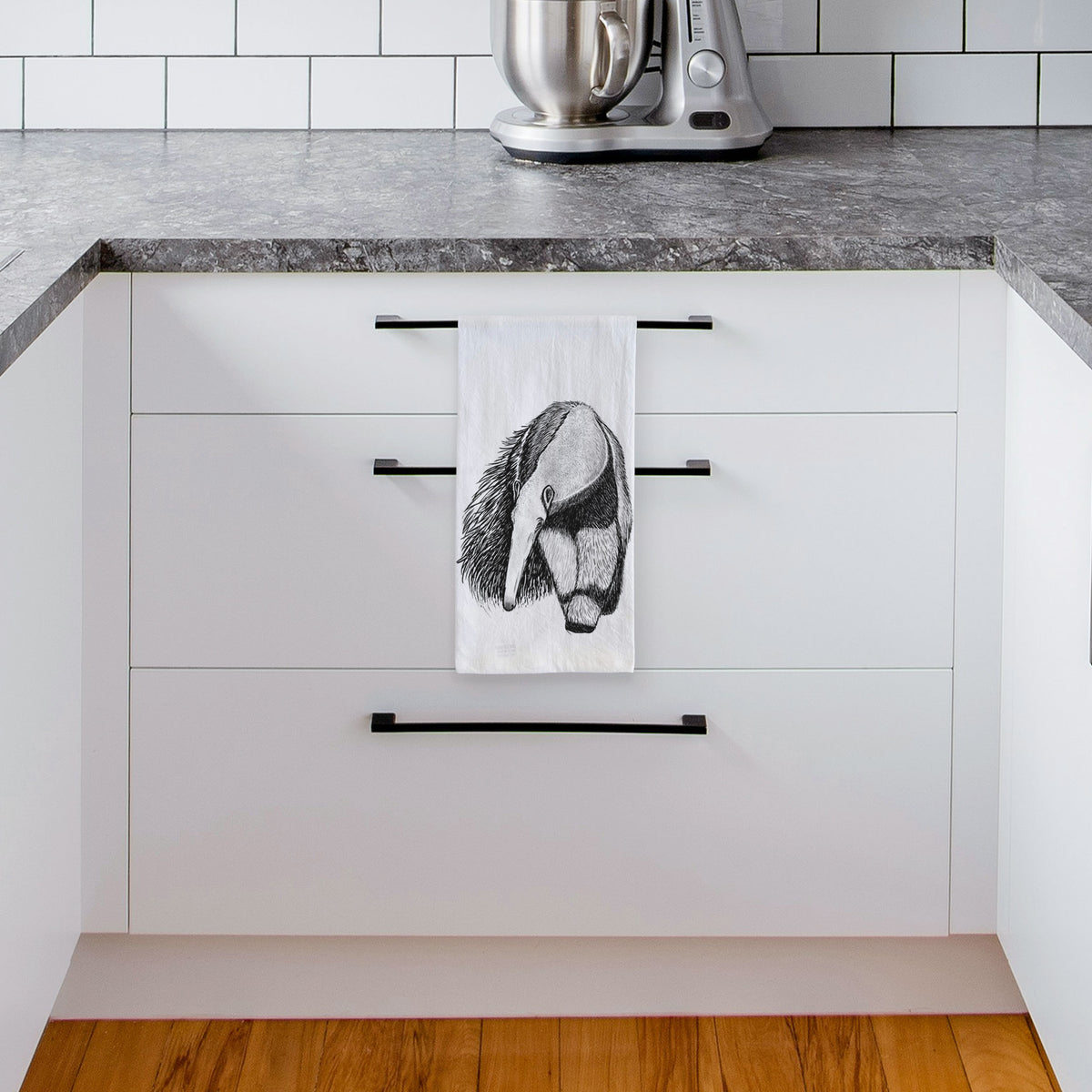 White tea towel printed with a detailed black ink illustration of a giant anteater with its long snout and bushy tail, hanging from a drawer handle in a bright white kitchen.