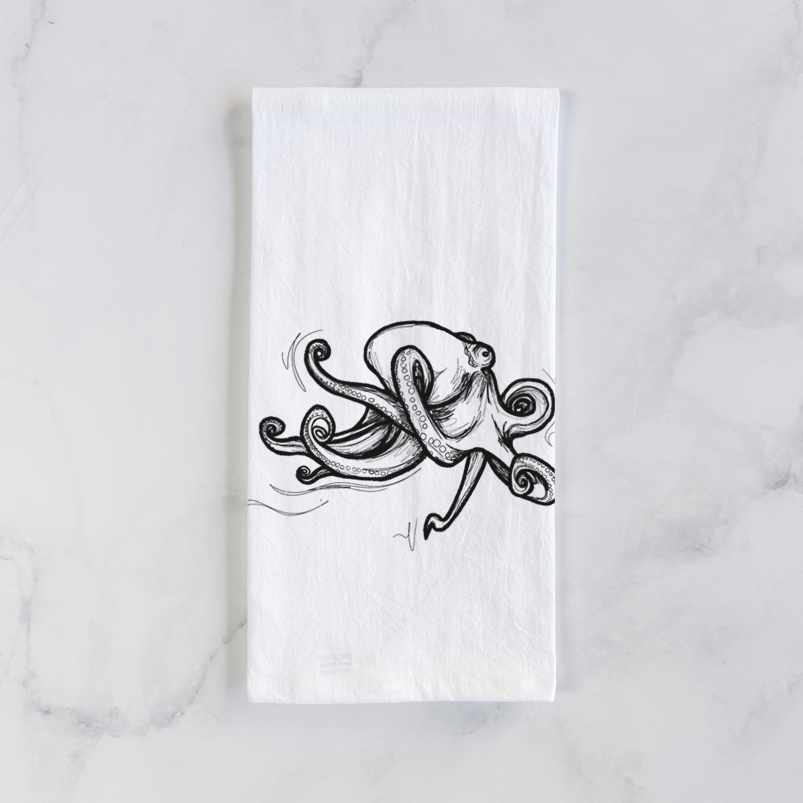 White cotton tea towel printed with a black ink illustration of a giant Pacific octopus with curling tentacles, on a marble counter beside a round wooden cutting board and greenery.