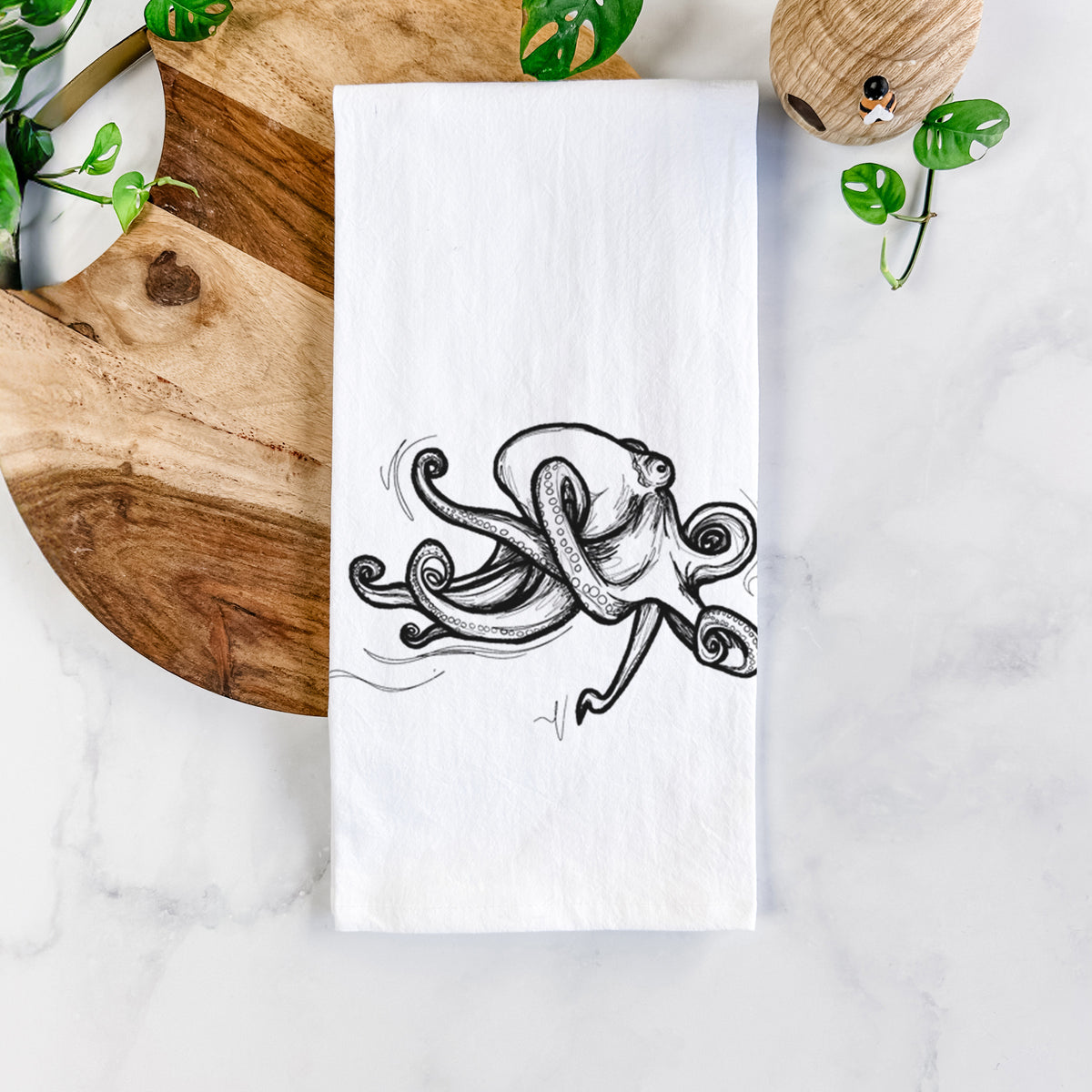 White cotton tea towel printed with a black ink illustration of a giant Pacific octopus with curling tentacles, on a marble counter beside a round wooden cutting board and greenery.