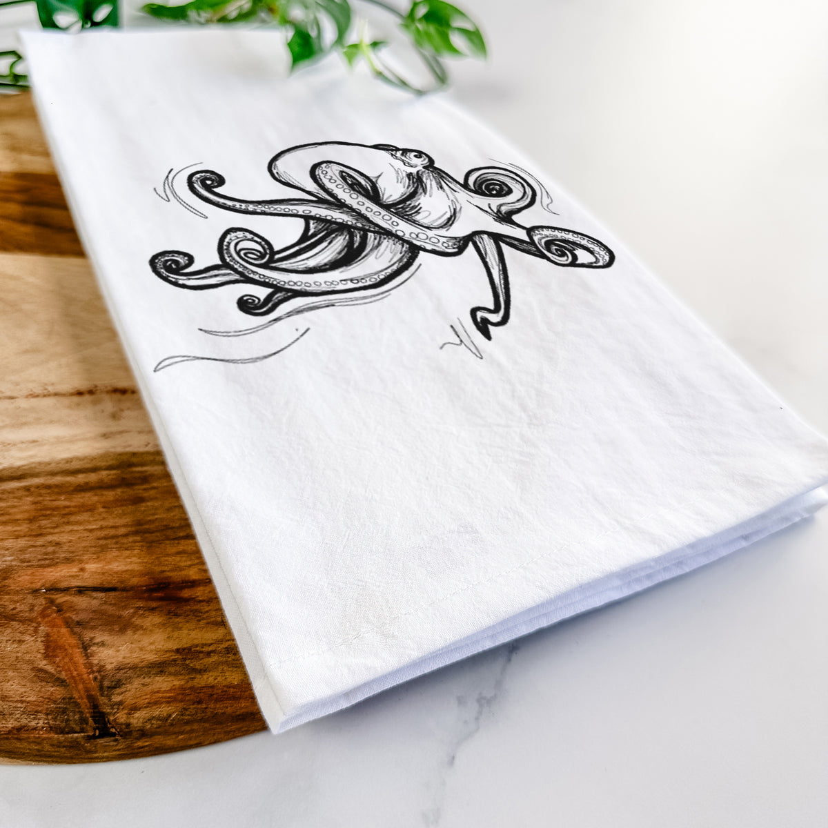 Close-up of a folded white tea towel on a wooden board, showing a black ink illustration of a giant Pacific octopus with curling tentacles.