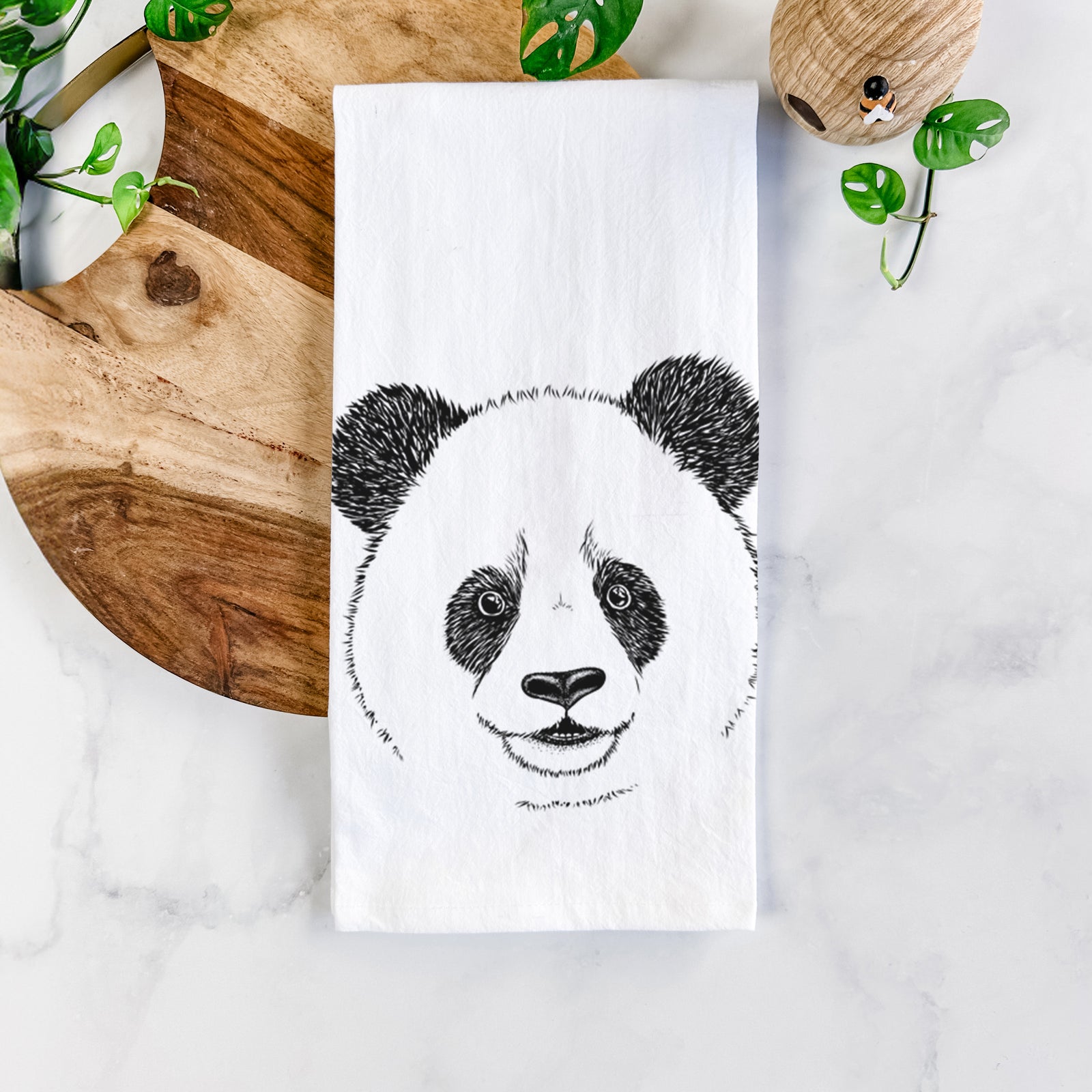 White cotton tea towel printed with a black ink portrait of a smiling giant panda's face, on a marble counter beside a round wooden cutting board and greenery.
