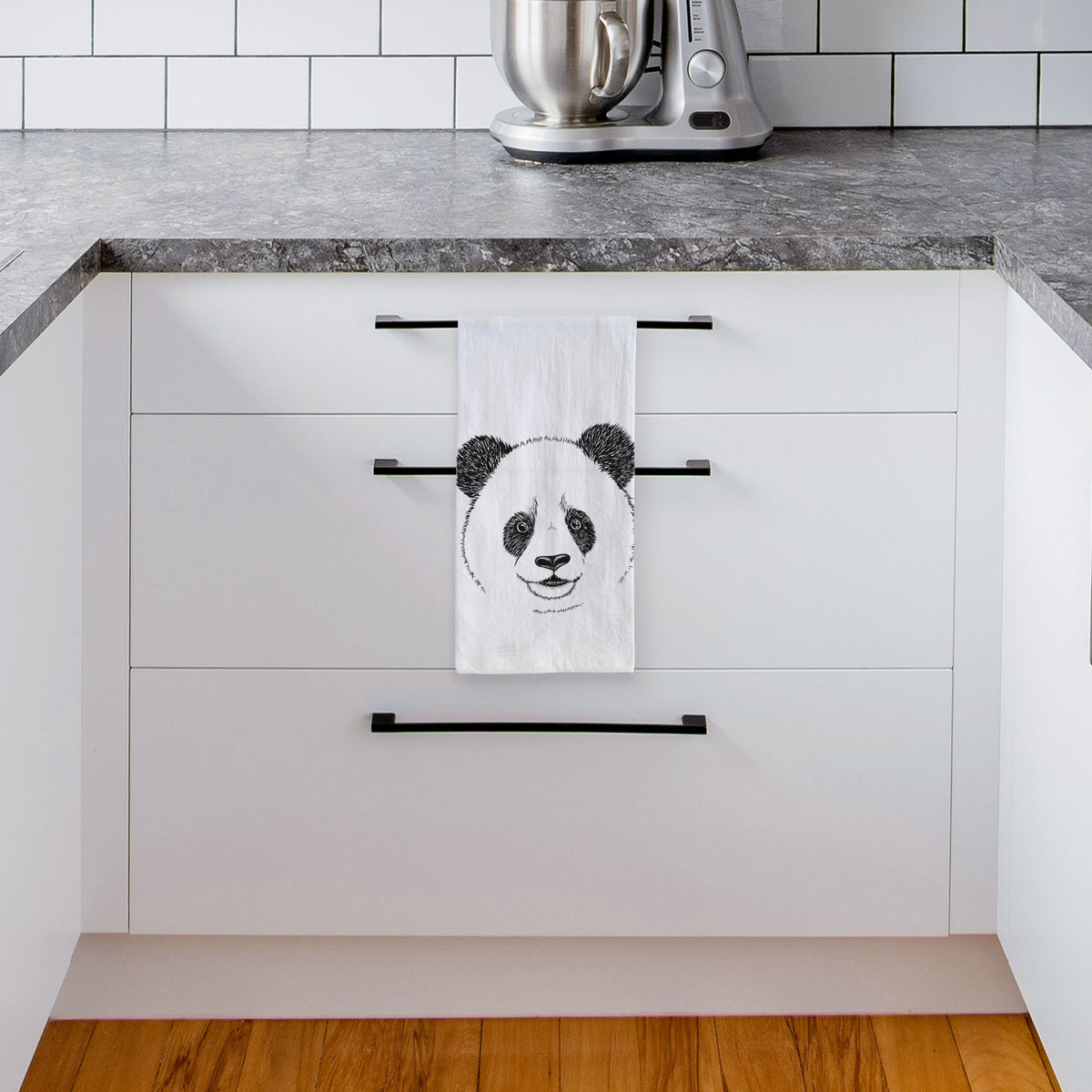White tea towel printed with a black ink portrait of a smiling giant panda's face, hanging from a drawer handle in a bright white kitchen.