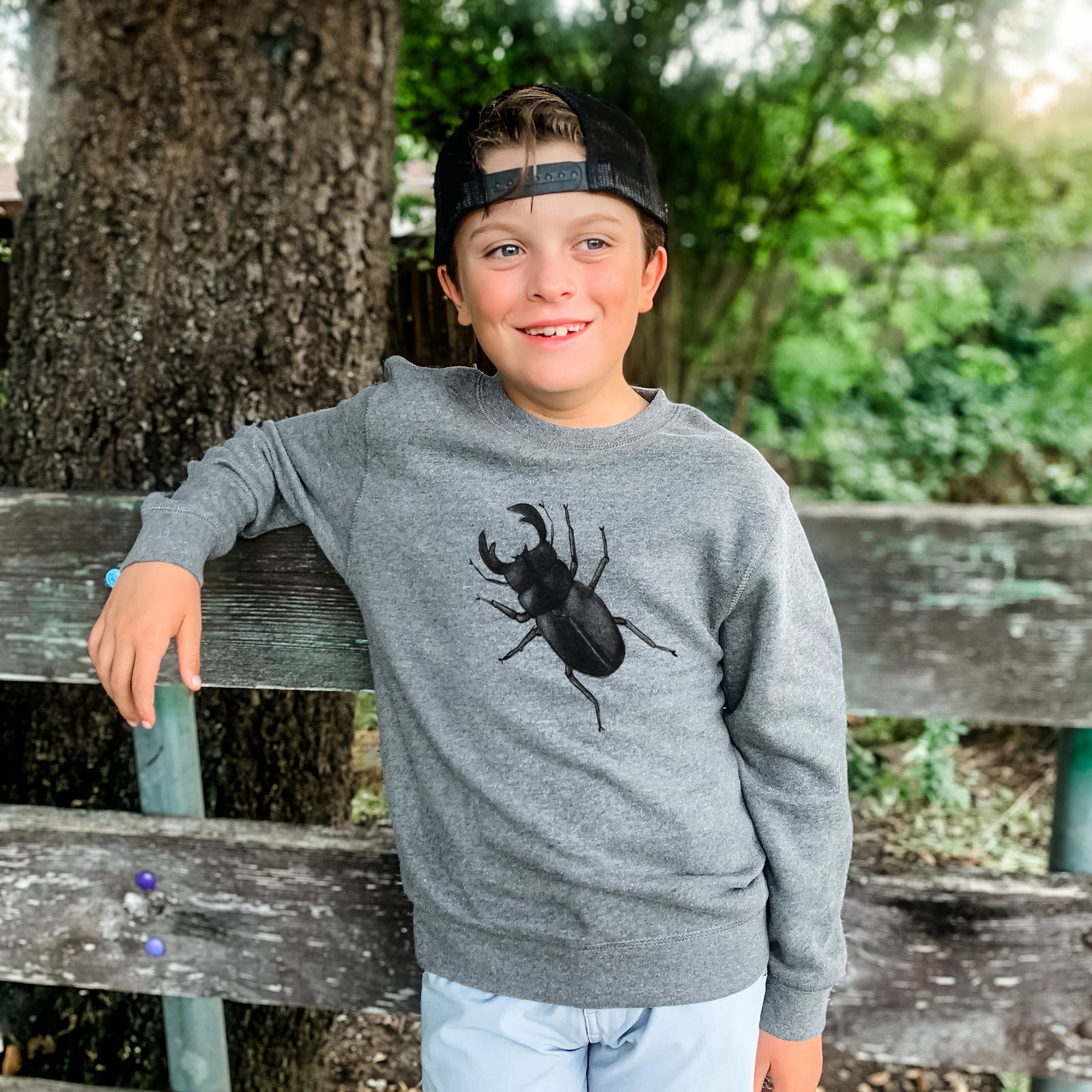 Boy wearing a youth crewneck sweatshirt printed with a detailed black ink illustration of a giant stag beetle with large jaws, seen from above, outdoors.