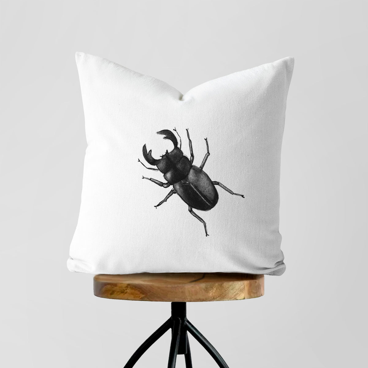 White pillow cover printed with a detailed black ink illustration of a giant stag beetle with large jaws, seen from above, on a wooden stool.