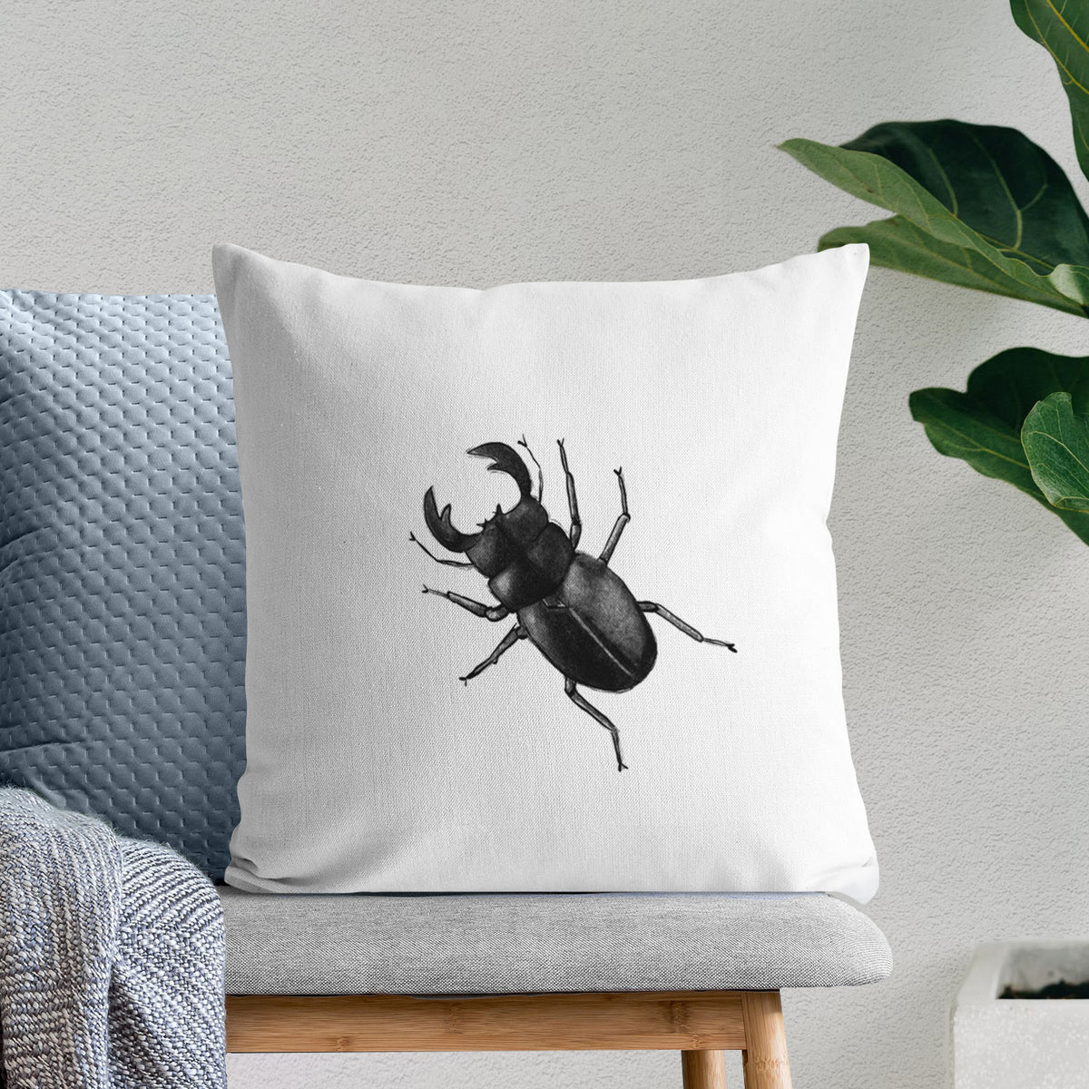 White pillow cover printed with a detailed black ink illustration of a giant stag beetle with large jaws, seen from above, on a bench beside plants.