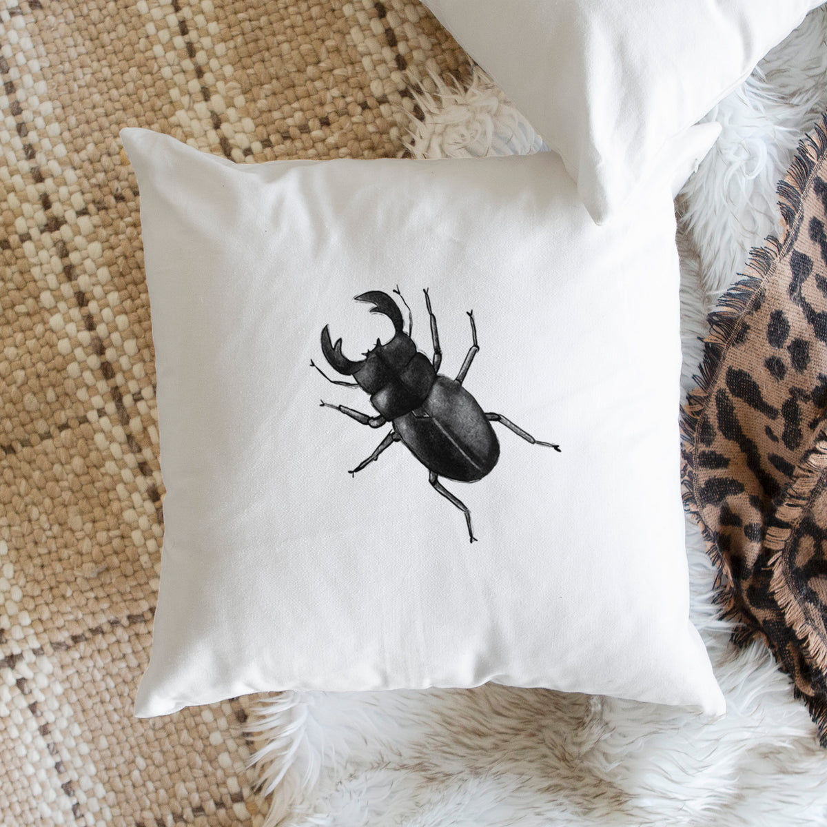 White pillow cover printed with a detailed black ink illustration of a giant stag beetle with large jaws, seen from above, on a woven chair.