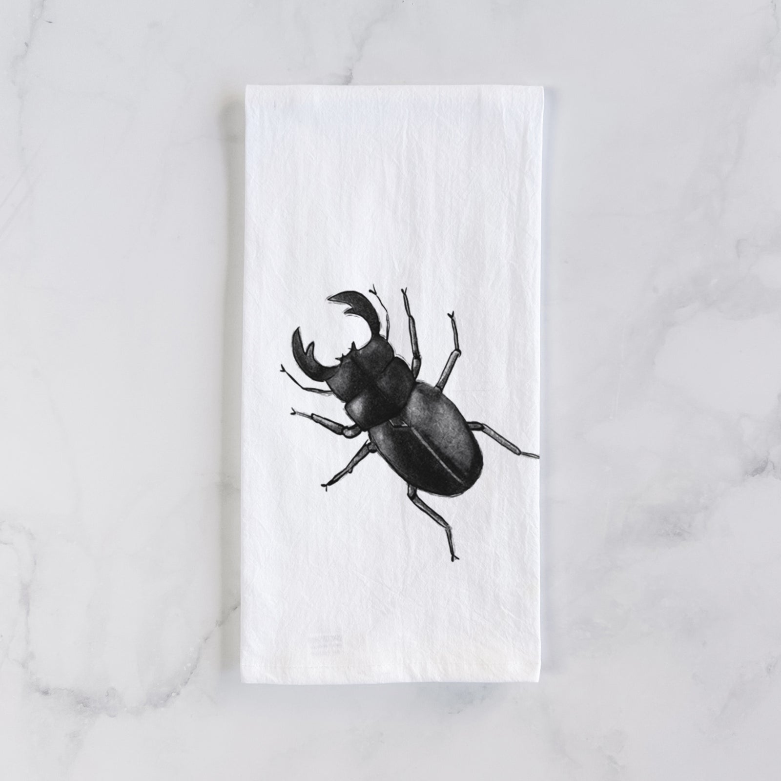 White cotton tea towel printed with a detailed black ink illustration of a giant stag beetle with large jaws, seen from above, on a marble counter beside a round wooden cutting board and greenery.