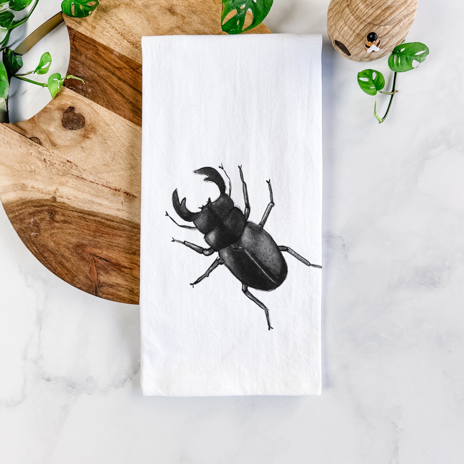 White cotton tea towel printed with a detailed black ink illustration of a giant stag beetle with large jaws, seen from above, on a marble counter beside a round wooden cutting board and greenery.