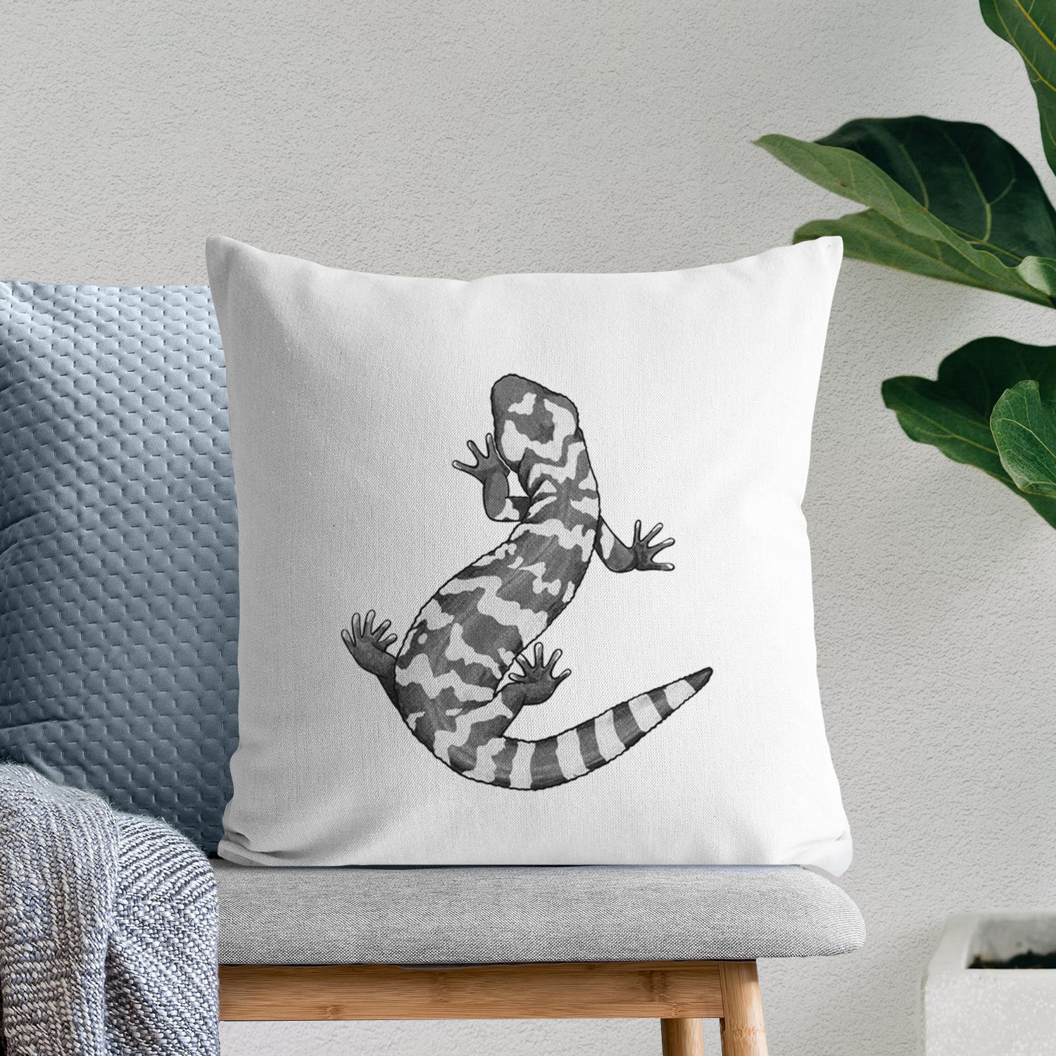 White pillow cover printed with a black ink illustration of a Gila monster with bold patterned skin, on a bench beside plants.