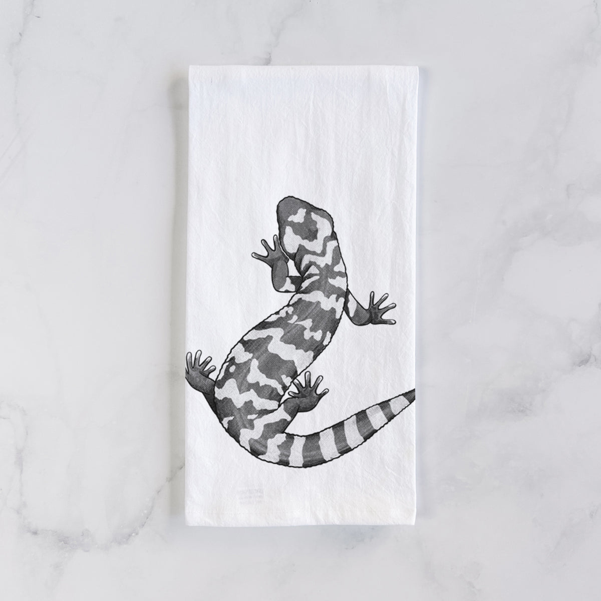 White cotton tea towel laid flat on marble, printed with a black ink illustration of a Gila monster with bold patterned skin.